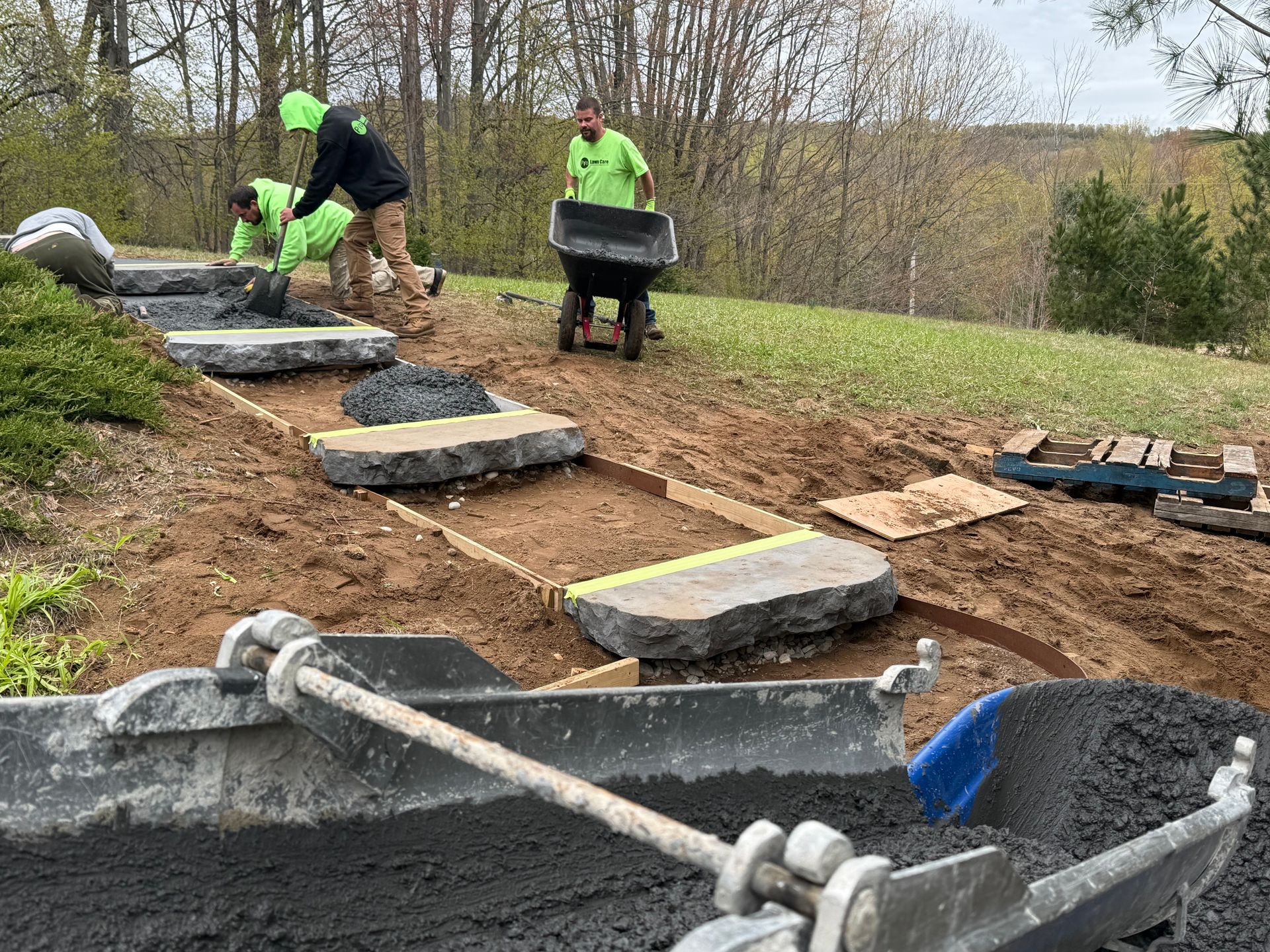 Workers in neon hats laying large stone slabs along a dirt path in a wooded area