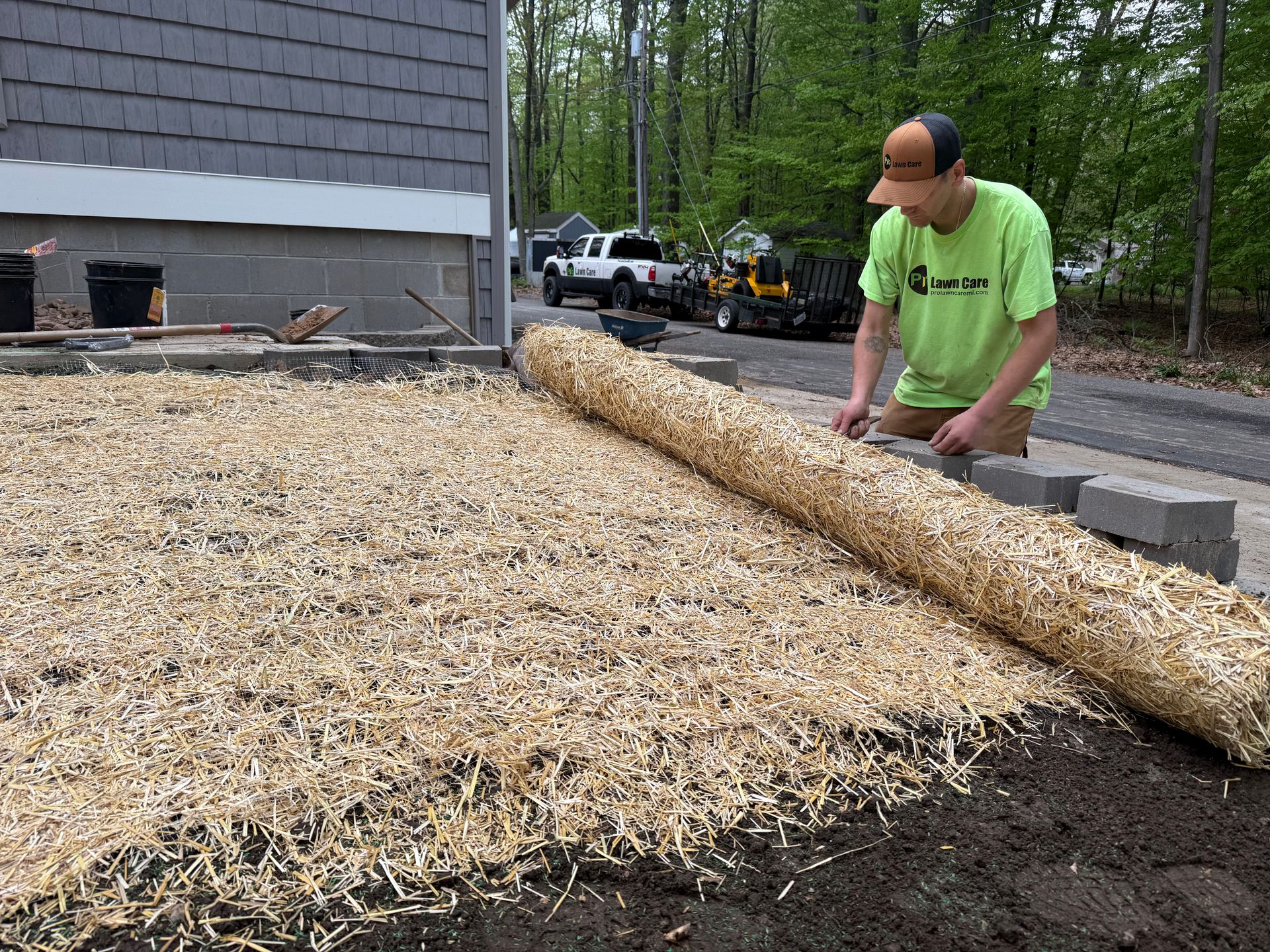 Person in neon shirt spreading gravel beside a house, with a long log and wooded yard nearby