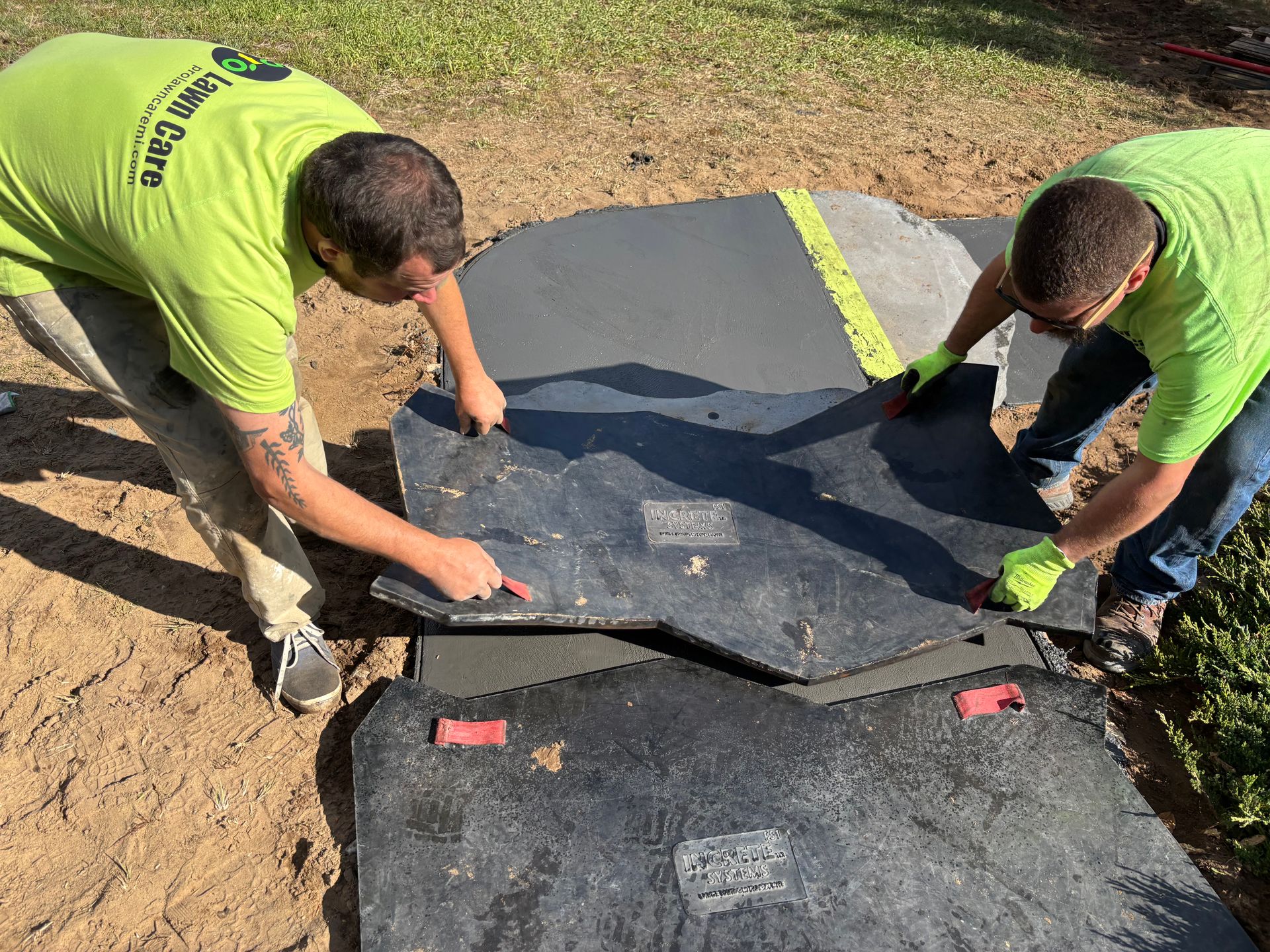 Two workers in green shirts fitting black paving stones onto a curved path edge outdoors