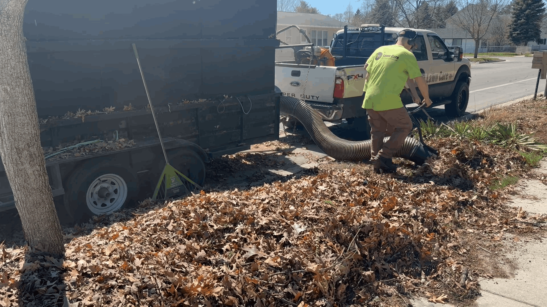 Worker in neon shirt shovels leaves beside a black trailer on a roadside in daylight