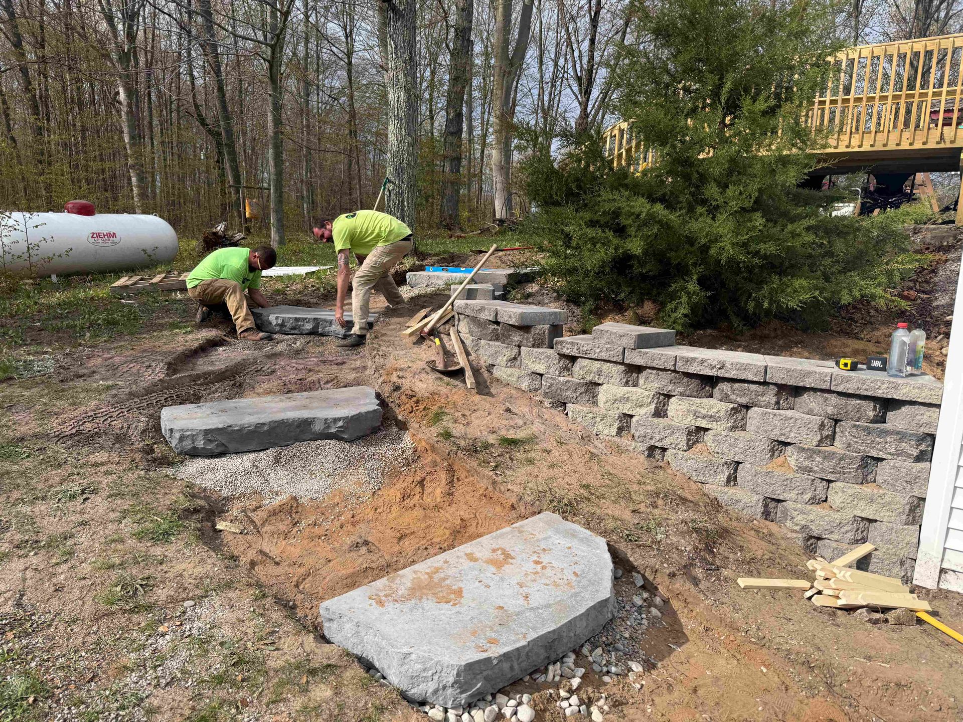 Workers building a stone retaining wall in a wooded yard, with large rocks and dirt steps.