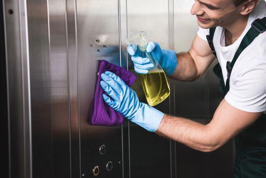 Persona che pulisce una superficie in acciaio inossidabile con un flacone spray e un panno viola, indossando guanti blu.
