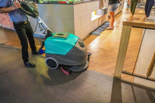 Una persona spinge una lavasciuga pavimenti color verde acqua in una corsia di un negozio, accanto a un bancone e a un tavolo.