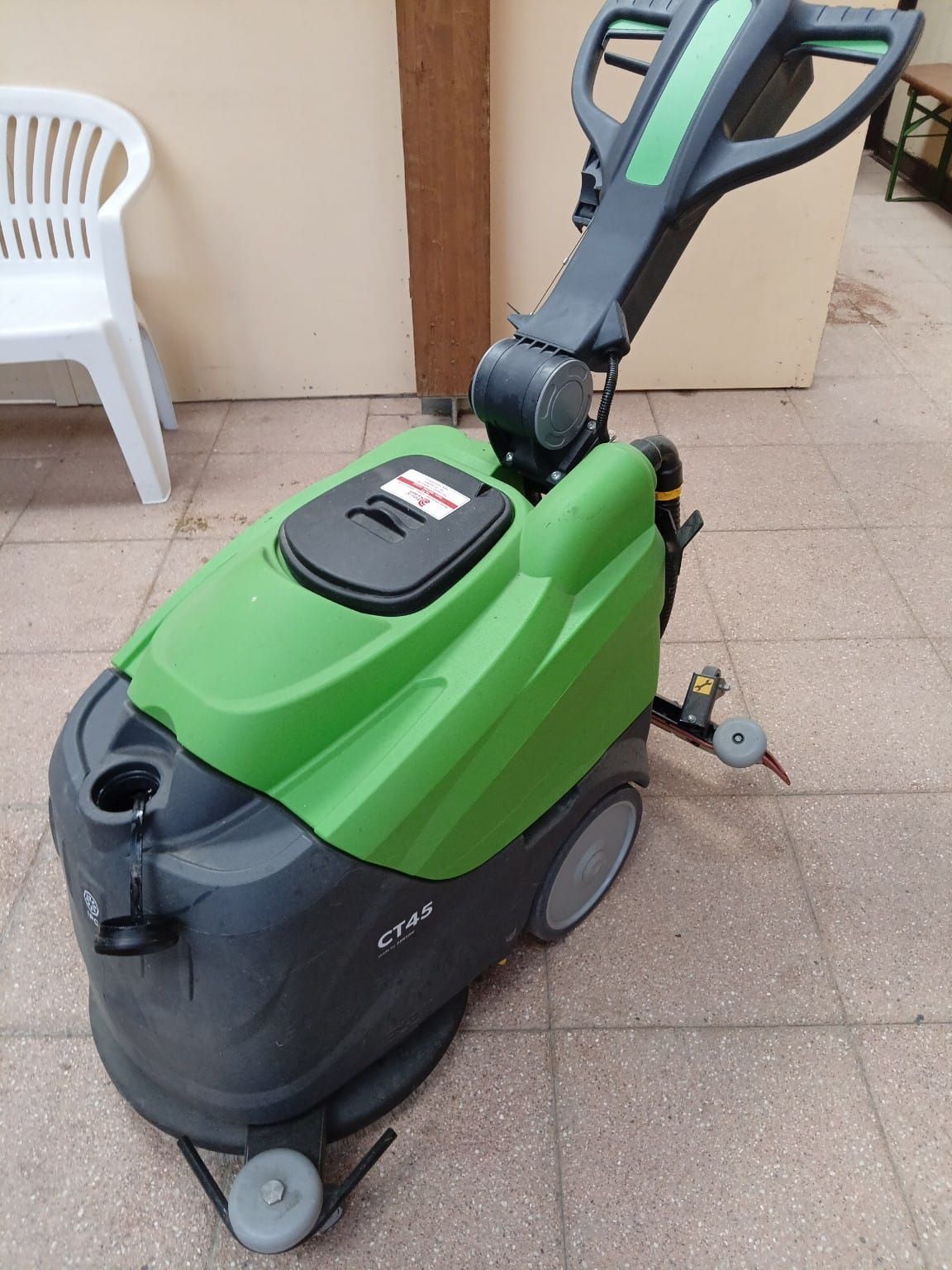 Una lavasciuga pavimenti verde con operatore a bordo è parcheggiata su un patio piastrellato, accanto a una sedia bianca e a una parete beige.
