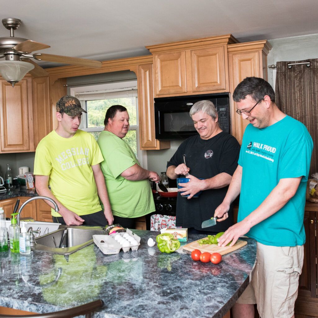 Four people in a kitchen preparing food. One cuts vegetables, another mixes ingredients, two watch.