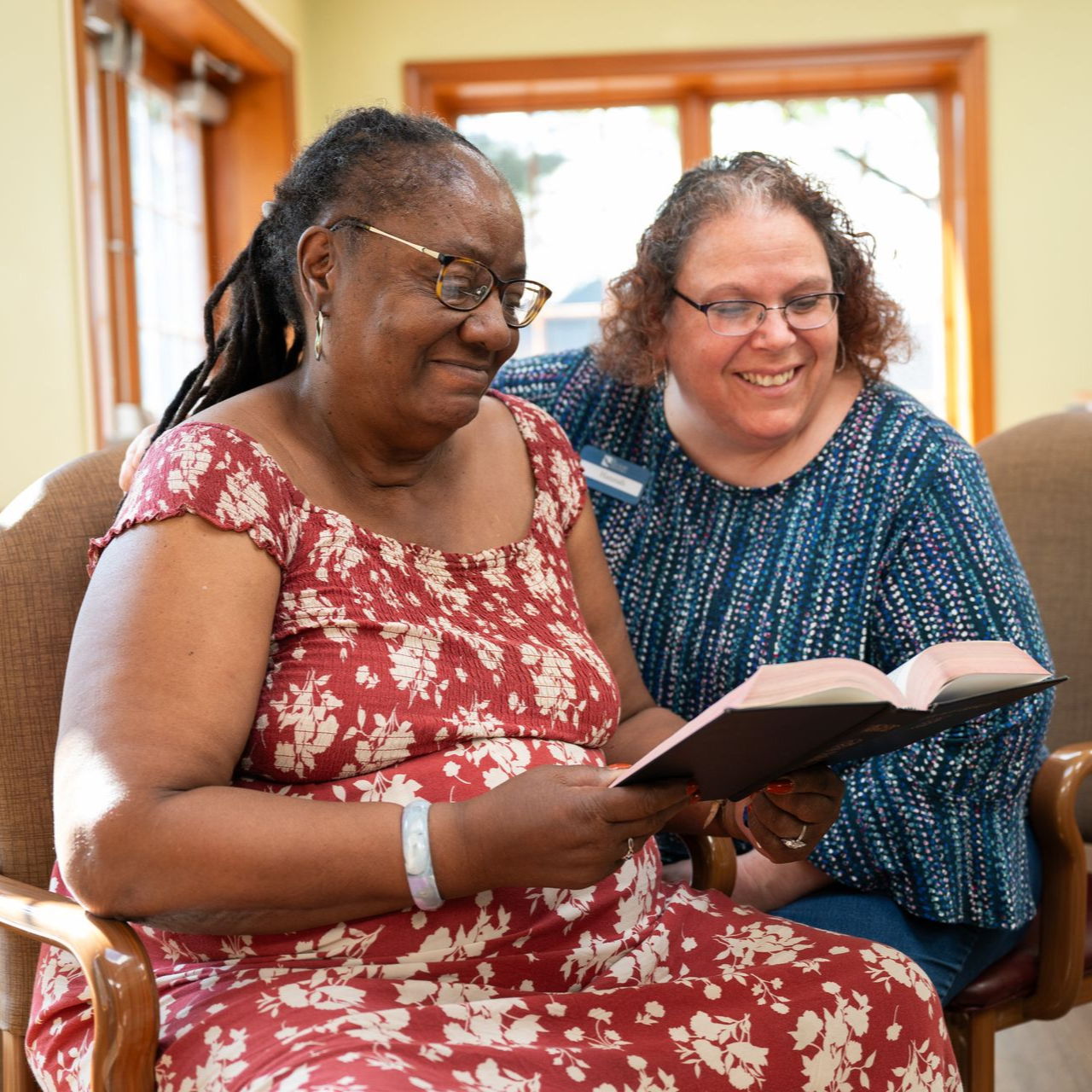 Two women reading a book, one with dreadlocks and red dress, the other in a blue top smiles.
