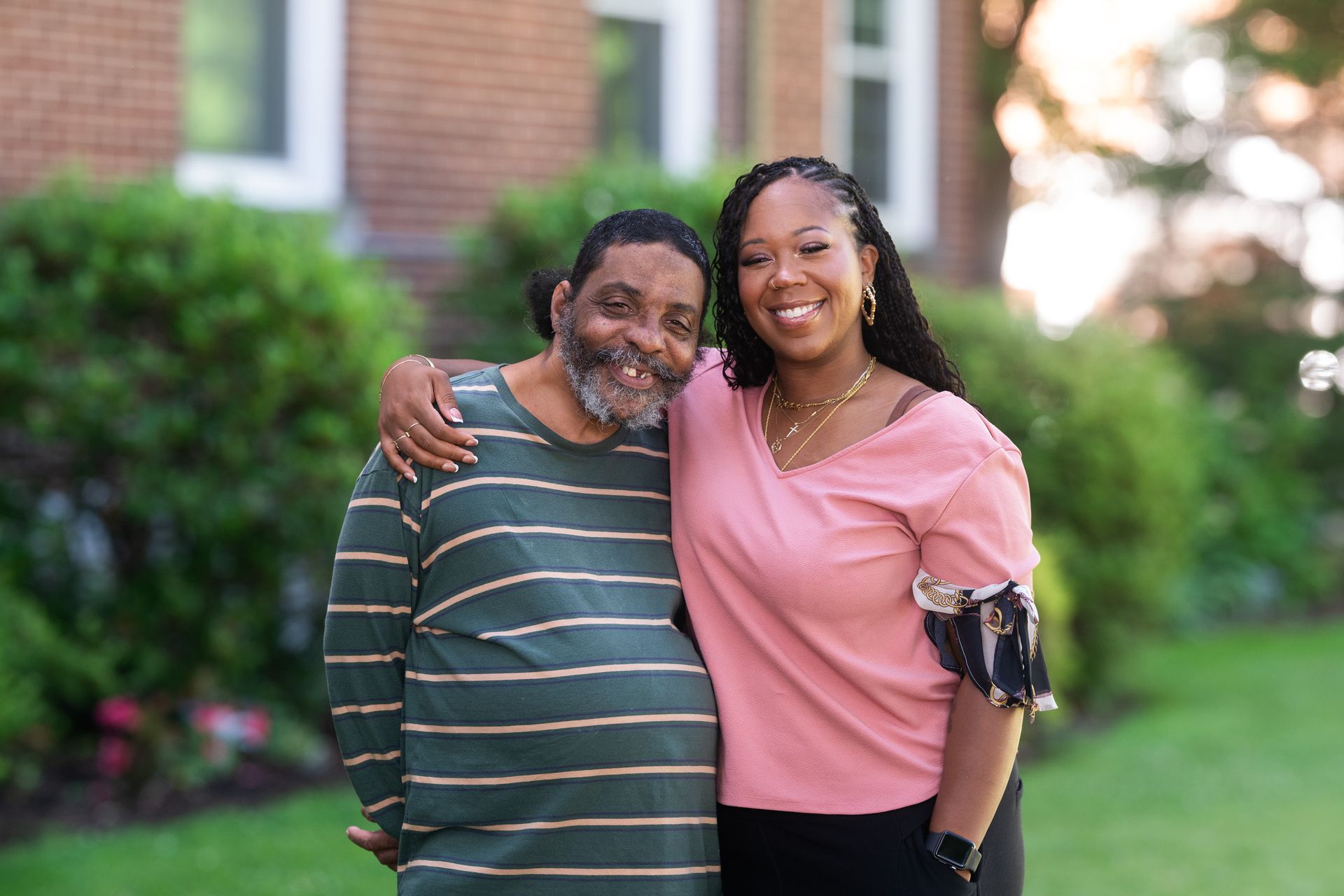 Two people smiling and embracing outdoors in a green garden, one in a striped shirt and one in a pink top