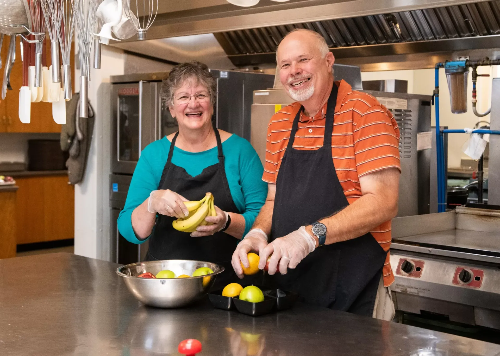 Two people preparing fruit in a commercial kitchen. Woman holds bananas, man cuts fruit.