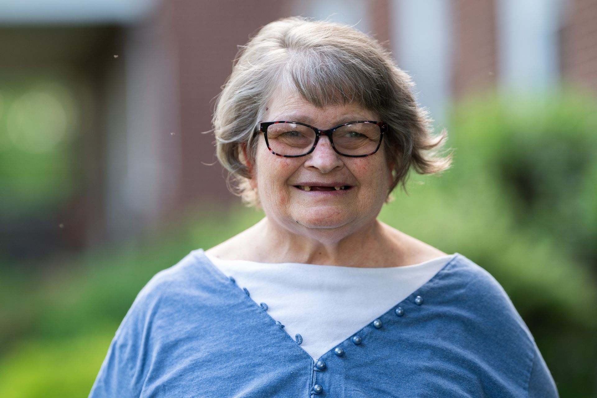 Woman with glasses, short gray hair, smiling, wearing a blue sweater over a white shirt, outdoors.