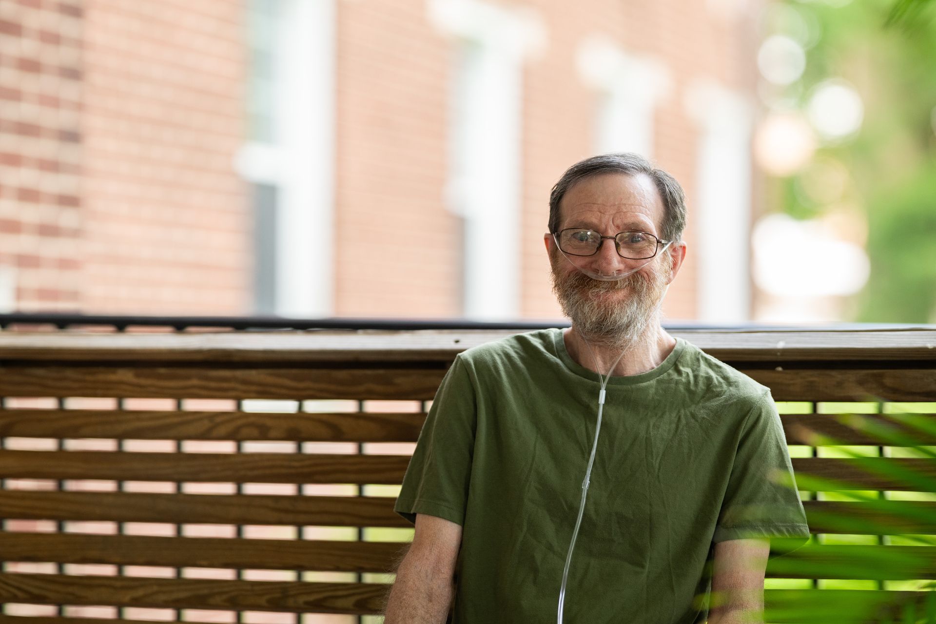 Man with glasses and oxygen tube smiling on a porch with wooden railing, brick building background.