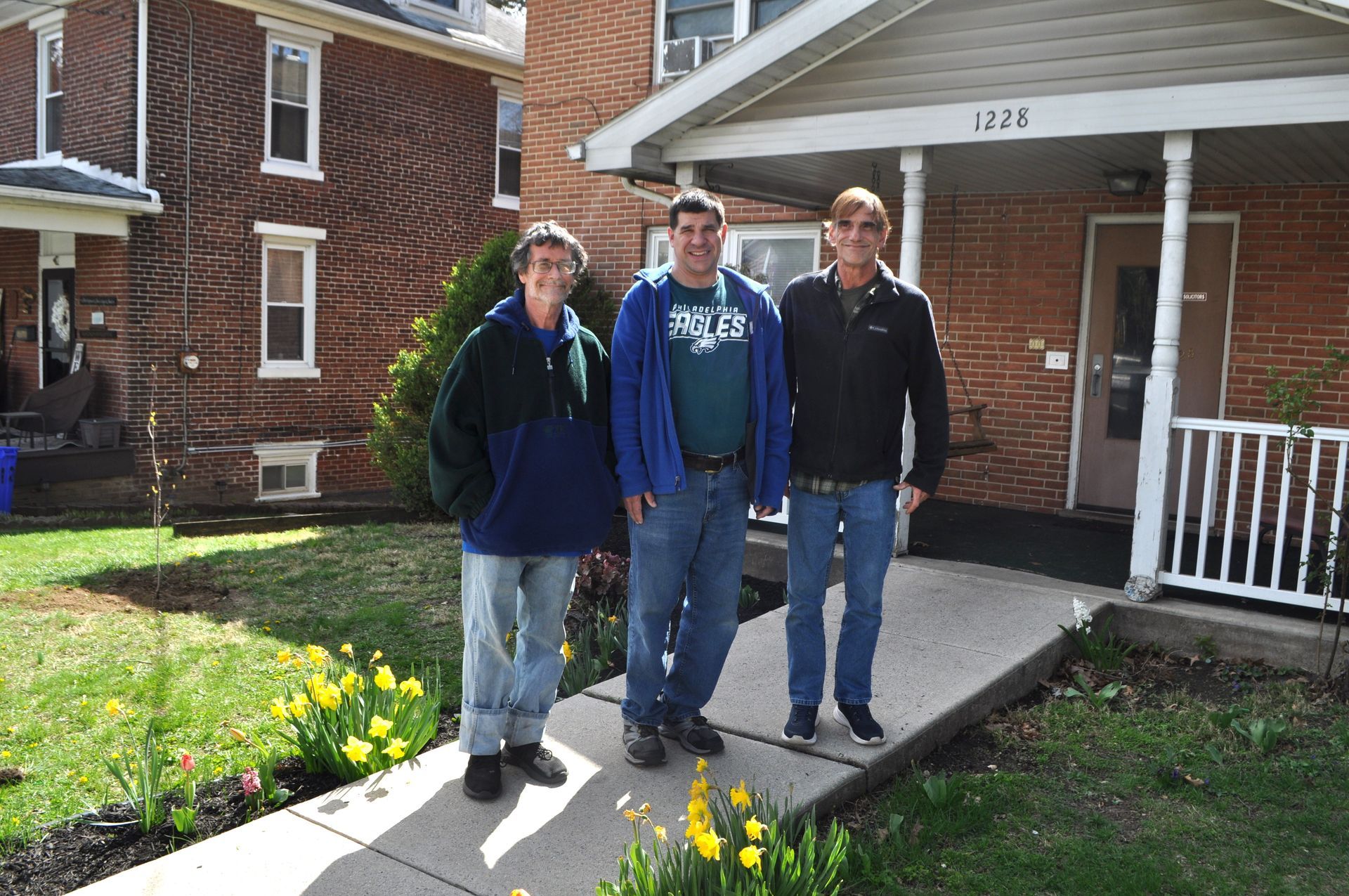 Three people standing in front of a brick house with a small porch, grass, and spring flowers.
