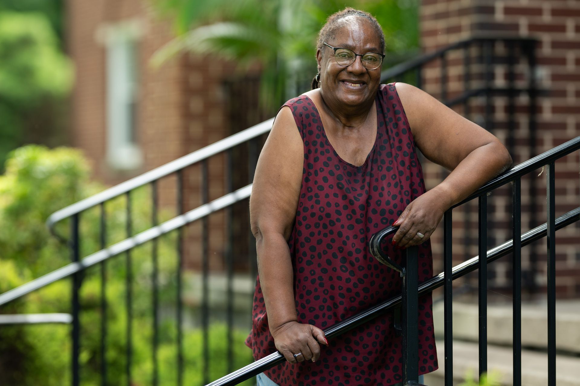 Woman in burgundy top smiles, leaning on a black railing. Outdoor setting with greenery and a brick building.