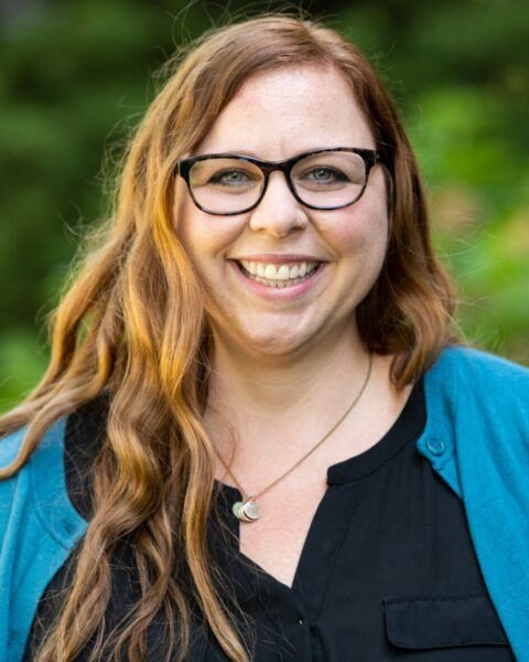 Woman with glasses smiling, wearing a blue cardigan and black shirt against a blurred green background. - Jessie