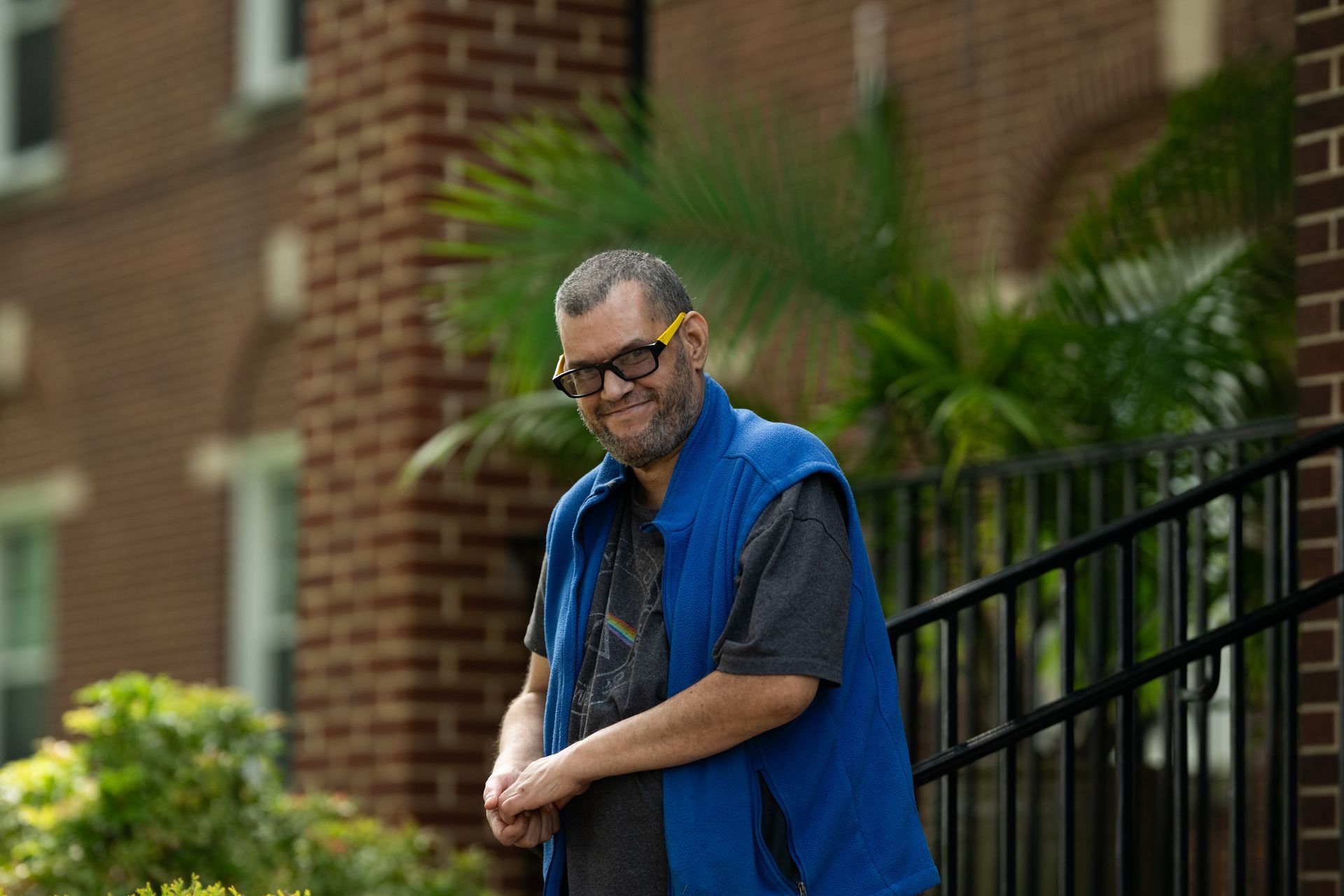 Man in blue vest and sunglasses smiles, standing near brick building.