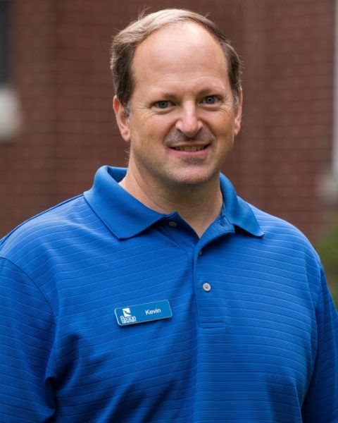 Man wearing a blue polo shirt, smiling, standing outdoors with a brick building in the background.