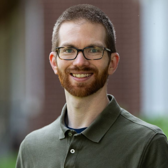 Man with a red beard and glasses smiles, wearing a green polo shirt, outside a brick building. - Andrew McNicol