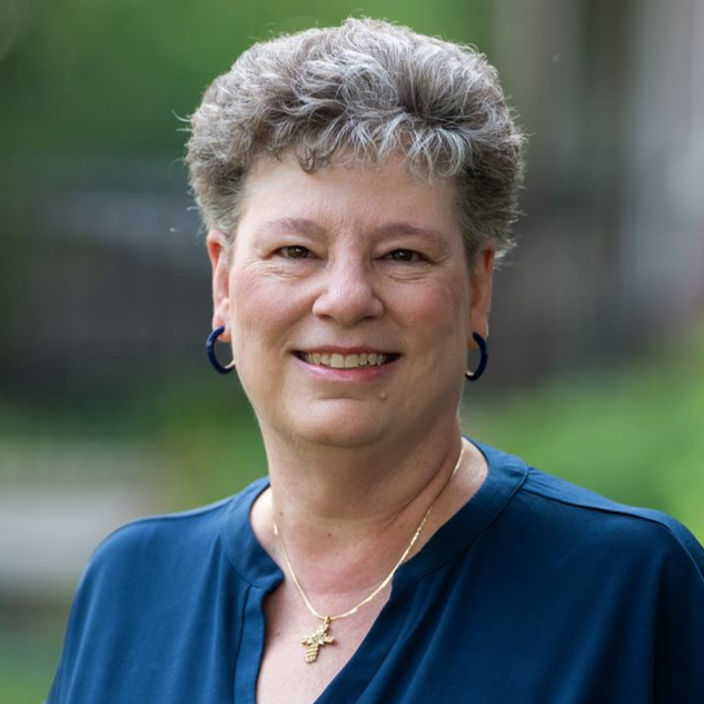 Woman with short gray hair smiles, wearing a blue shirt and gold cross necklace; outdoors. - Marla Sheaffer