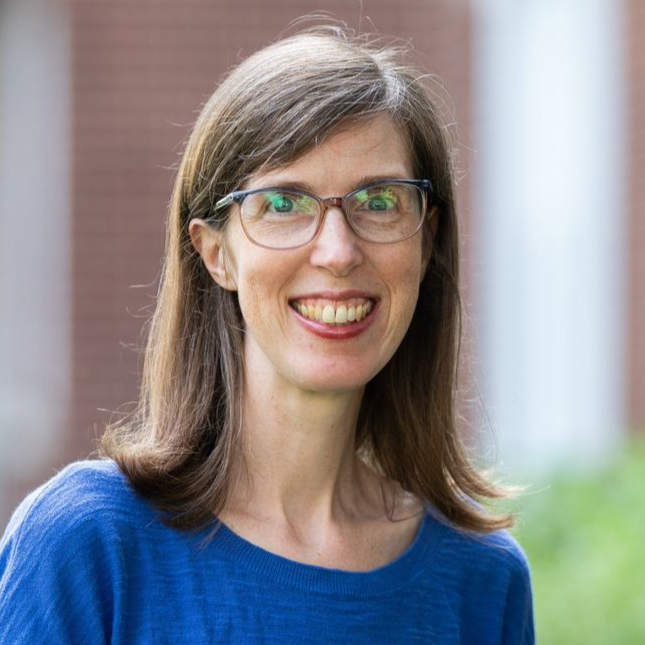 Woman with glasses smiling, wearing blue sweater, in front of a brick building. - Rachel Keen