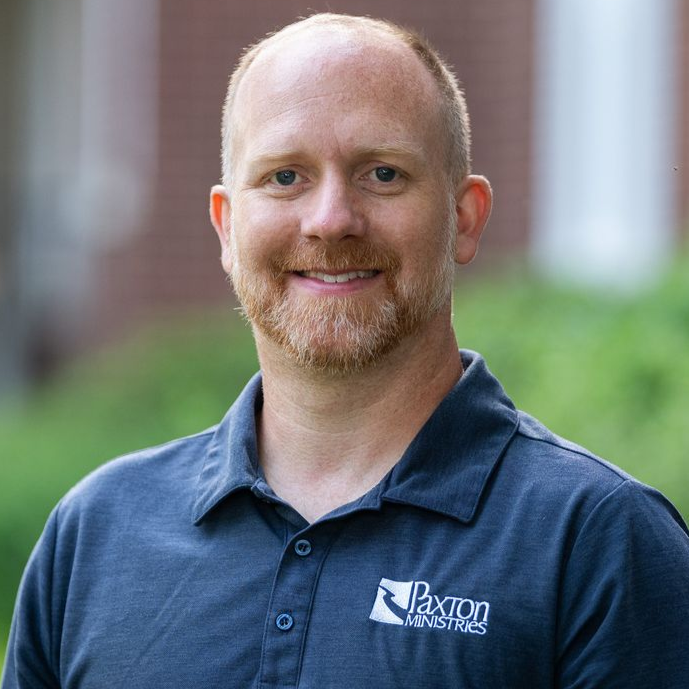 Man with reddish beard and polo shirt smiling outdoors. The shirt has a