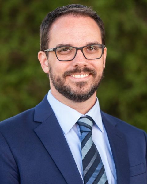 Man with glasses, beard, suit, and tie smiling outdoors in front of green foliage.