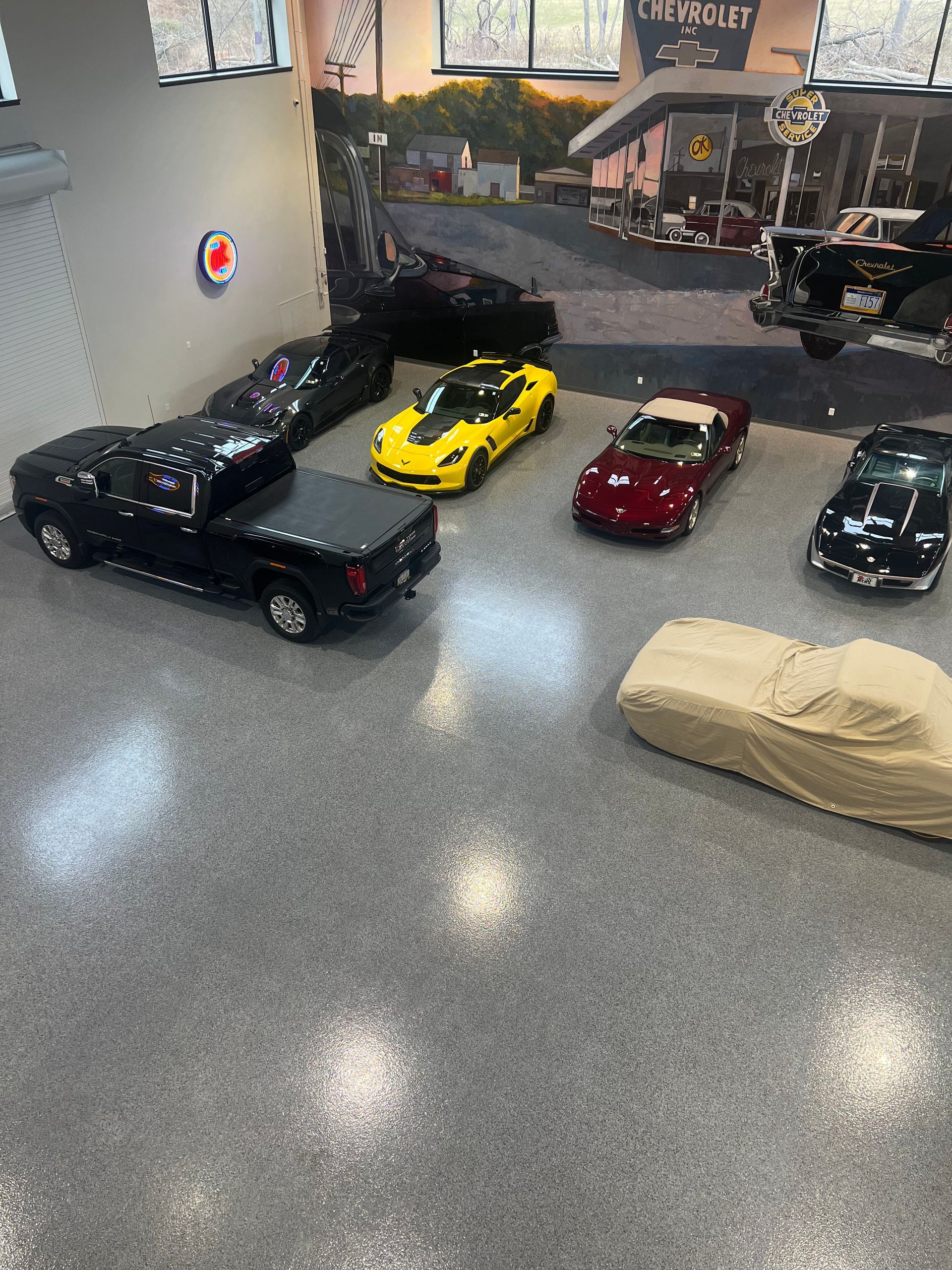 A high-angle view of a large, well-lit showroom featuring a black pickup truck, several sports cars, and a covered vehicle.