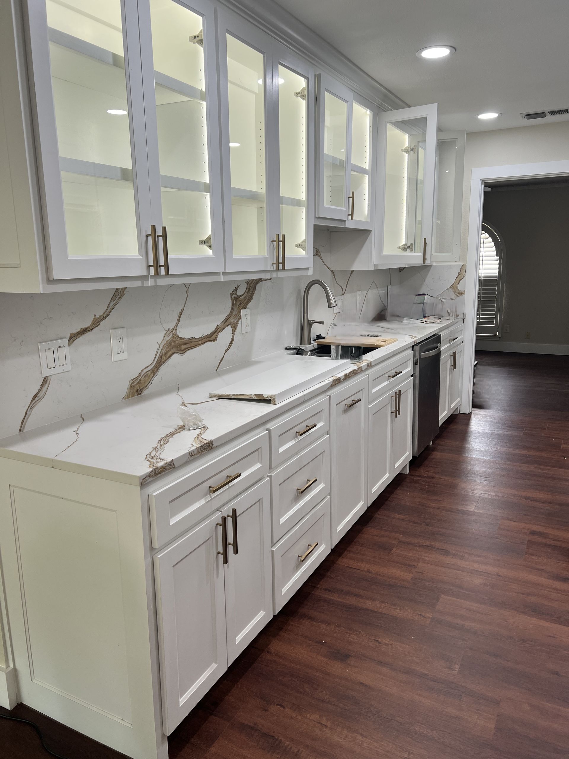 White kitchen cabinets with gold-veined countertops and glass-front uppers; view to living room.