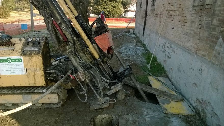 Una piattaforma di perforazione è posizionata accanto a un muro di mattoni, con un operaio in giacca arancione in piedi vicino all'attrezzatura.