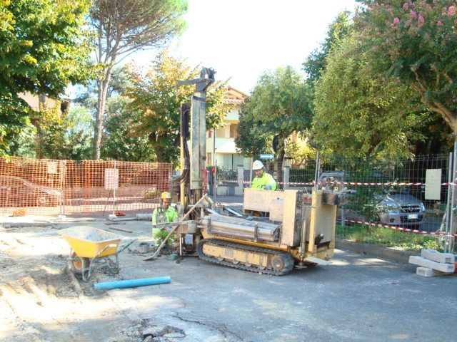 Un operaio edile manovra un piccolo escavatore in un cantiere stradale con alberi e recinzioni nelle vicinanze.