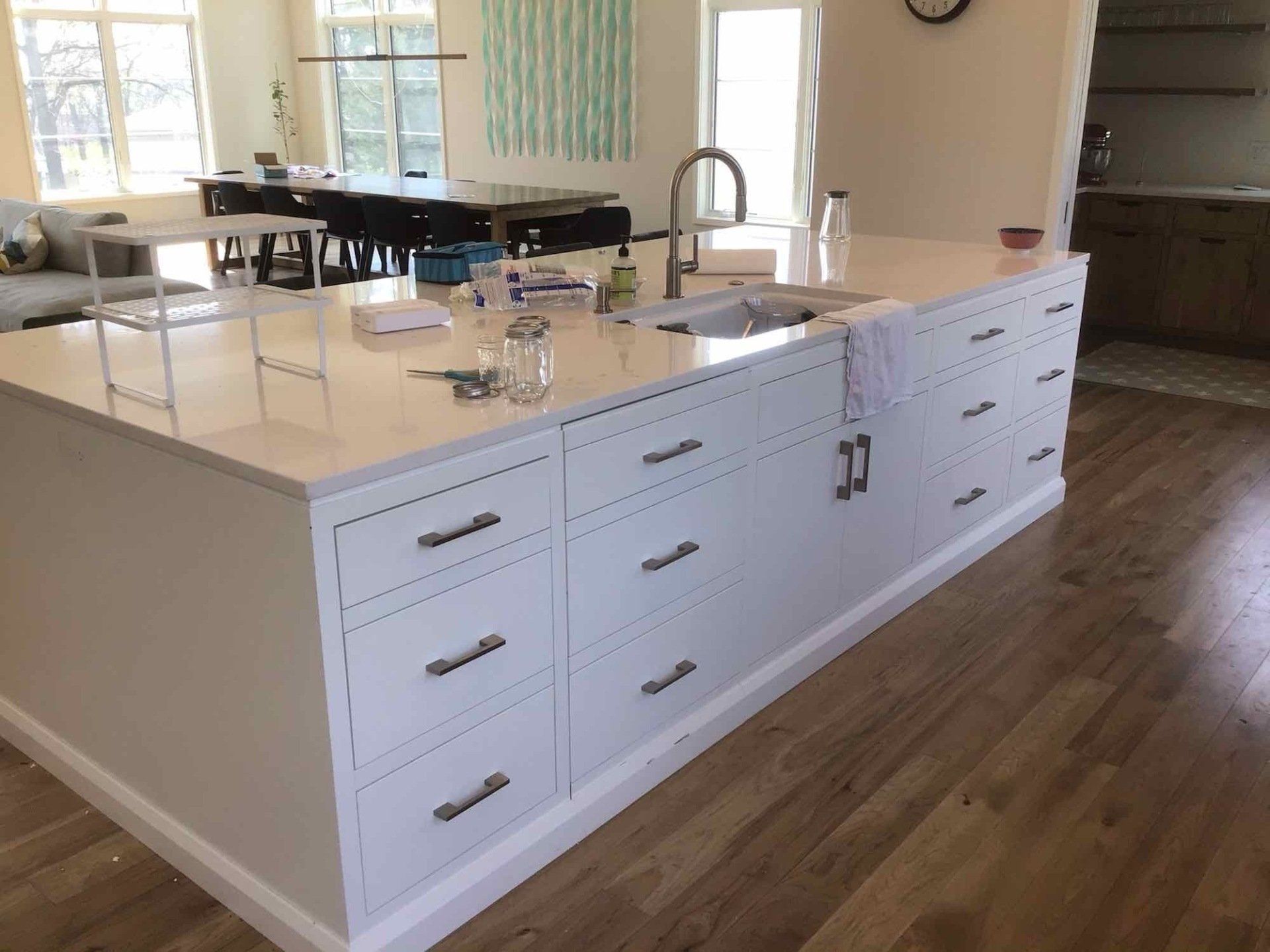 White kitchen island with drawers and sink, hardwood floor, light beige walls, and dining area in the background.