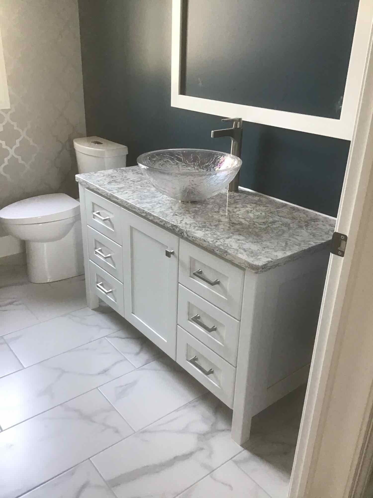 White bathroom vanity with granite countertop and glass sink. Toilet and mirror in the background.
