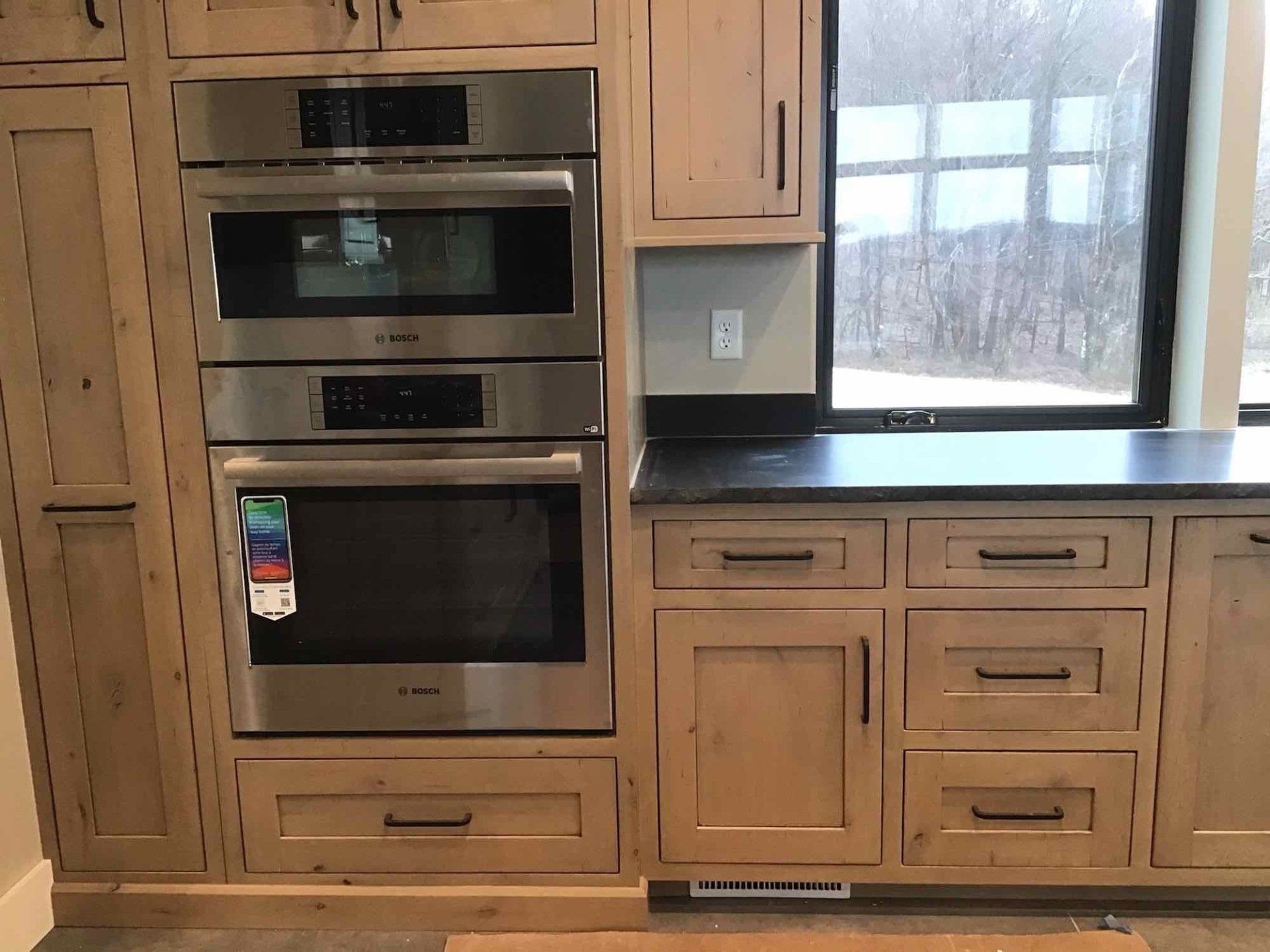 Stainless steel double ovens built into light wood cabinetry, with dark countertop and window.