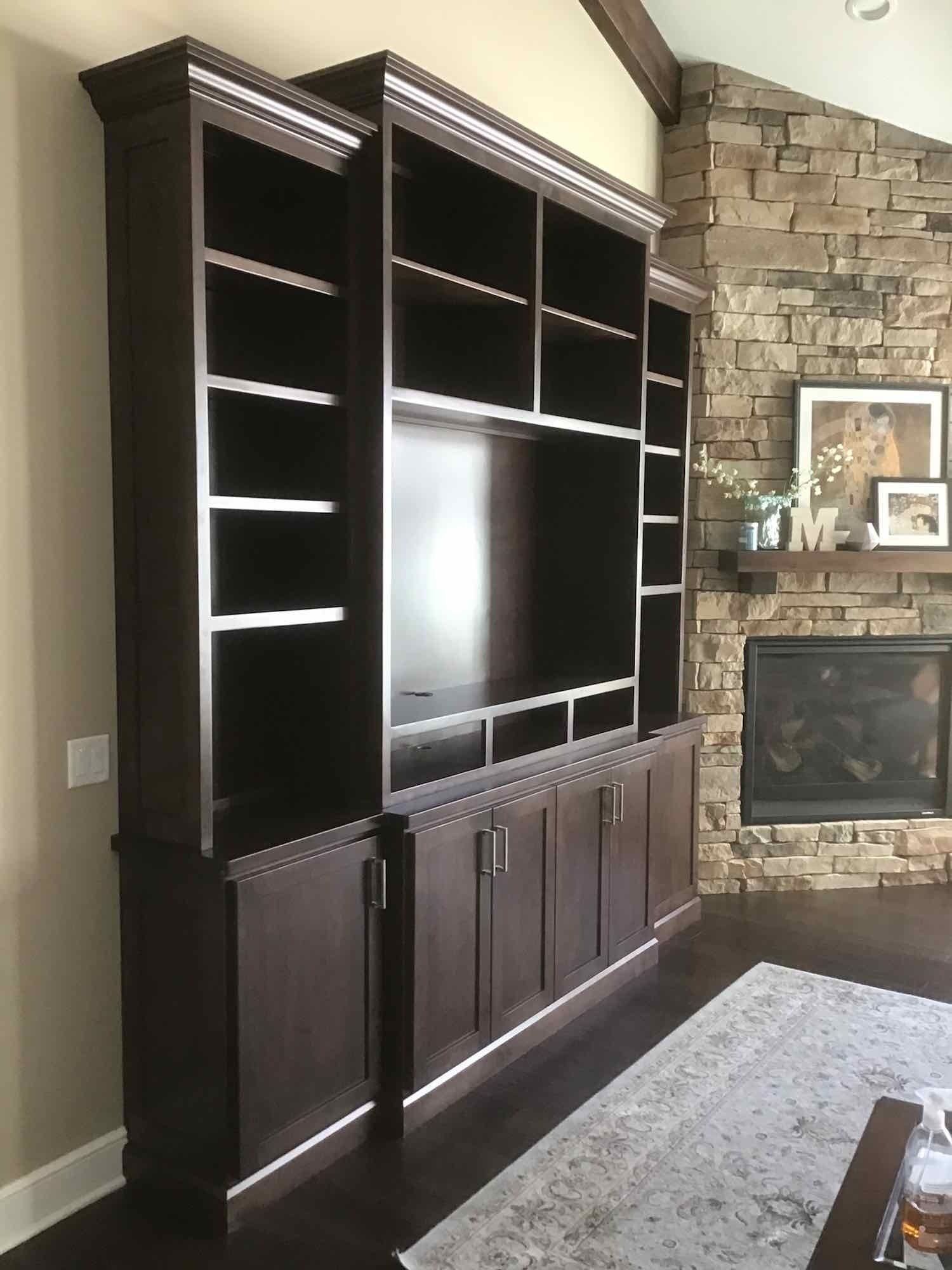 Dark brown built-in entertainment center with shelves, cabinets, and a television, next to a stone fireplace.