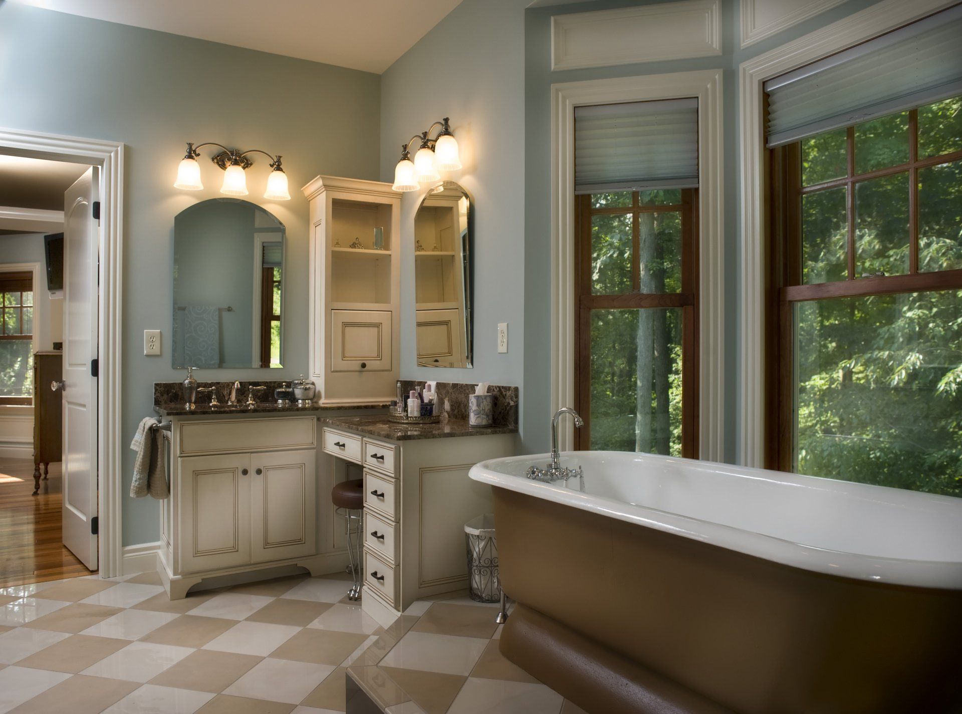 Bathroom with a freestanding bathtub, vanity with mirrors, and checkered floor. Windows offer a view of greenery.