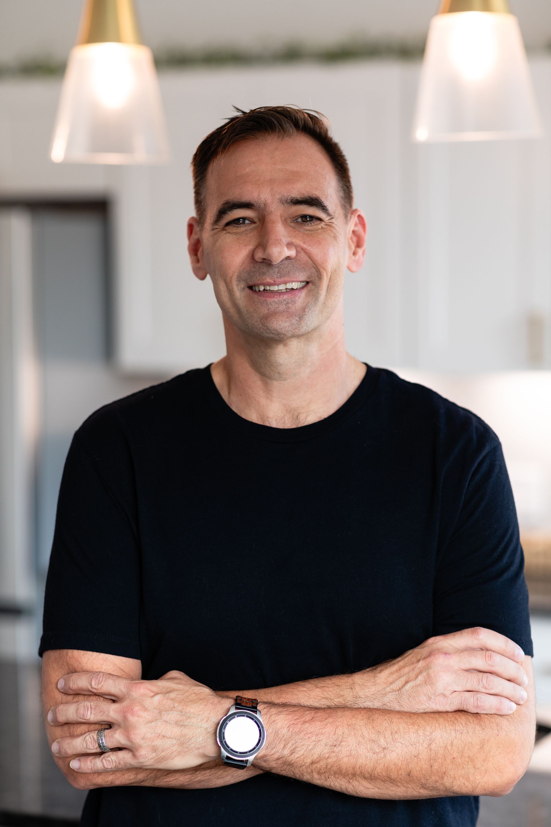 A man in a black shirt is standing with his arms crossed in a kitchen smiling.