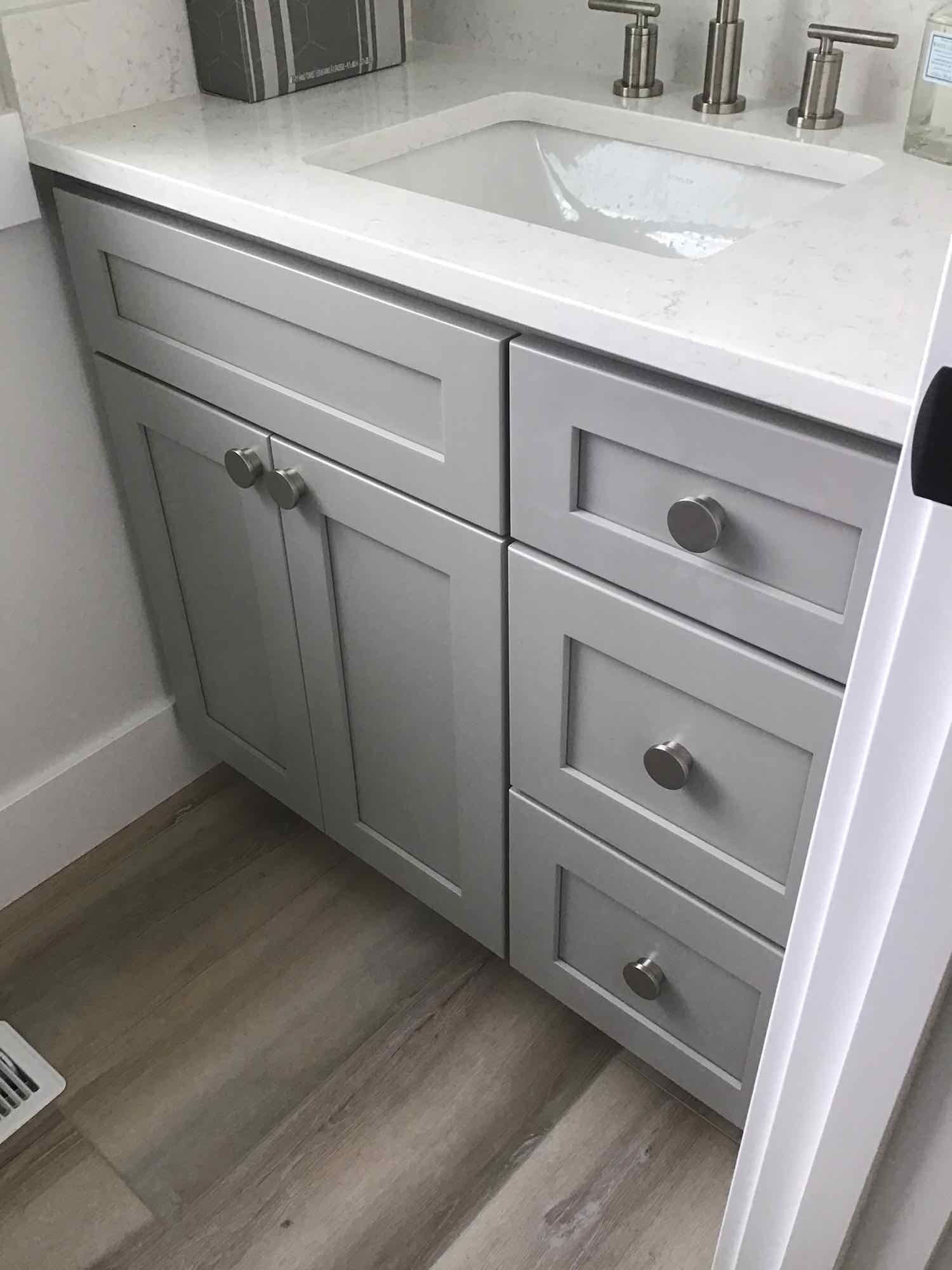 Gray bathroom vanity with white countertop and sink. Three drawers and a cabinet with silver hardware.
