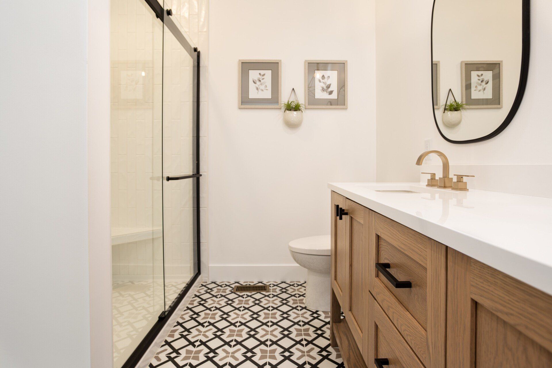 Bathroom with patterned floor, glass shower, light wood vanity, and round mirror.