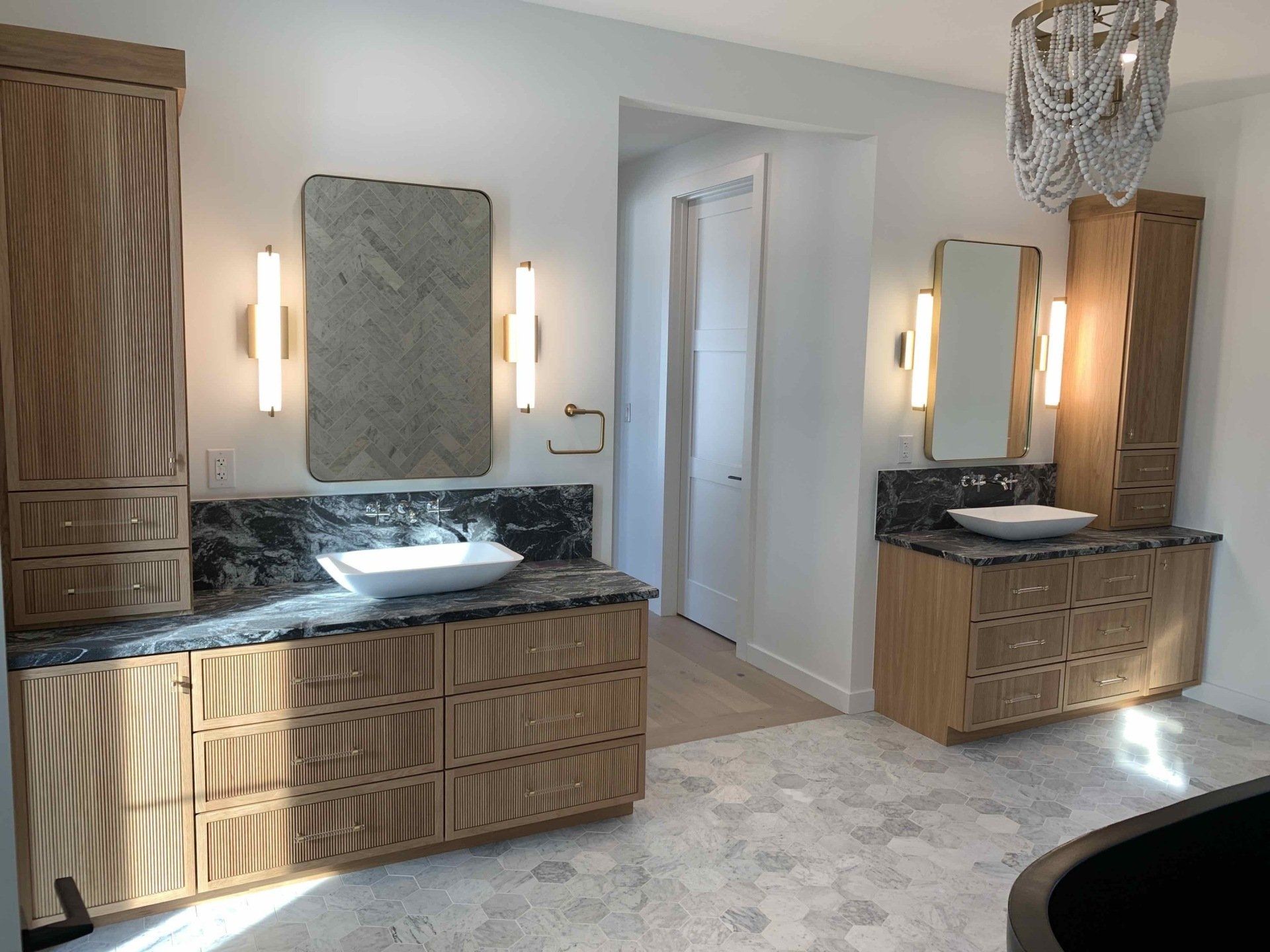 Two bathroom vanities with black countertops, light wood cabinets, mirrors, and a chandelier.