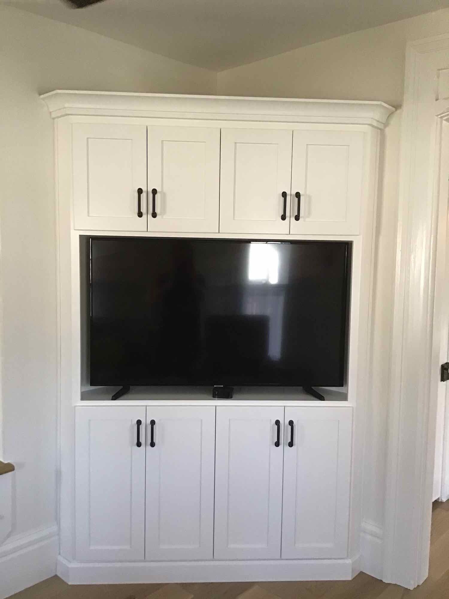White built-in cabinet with a black TV, in a corner. Cabinets above and below TV.