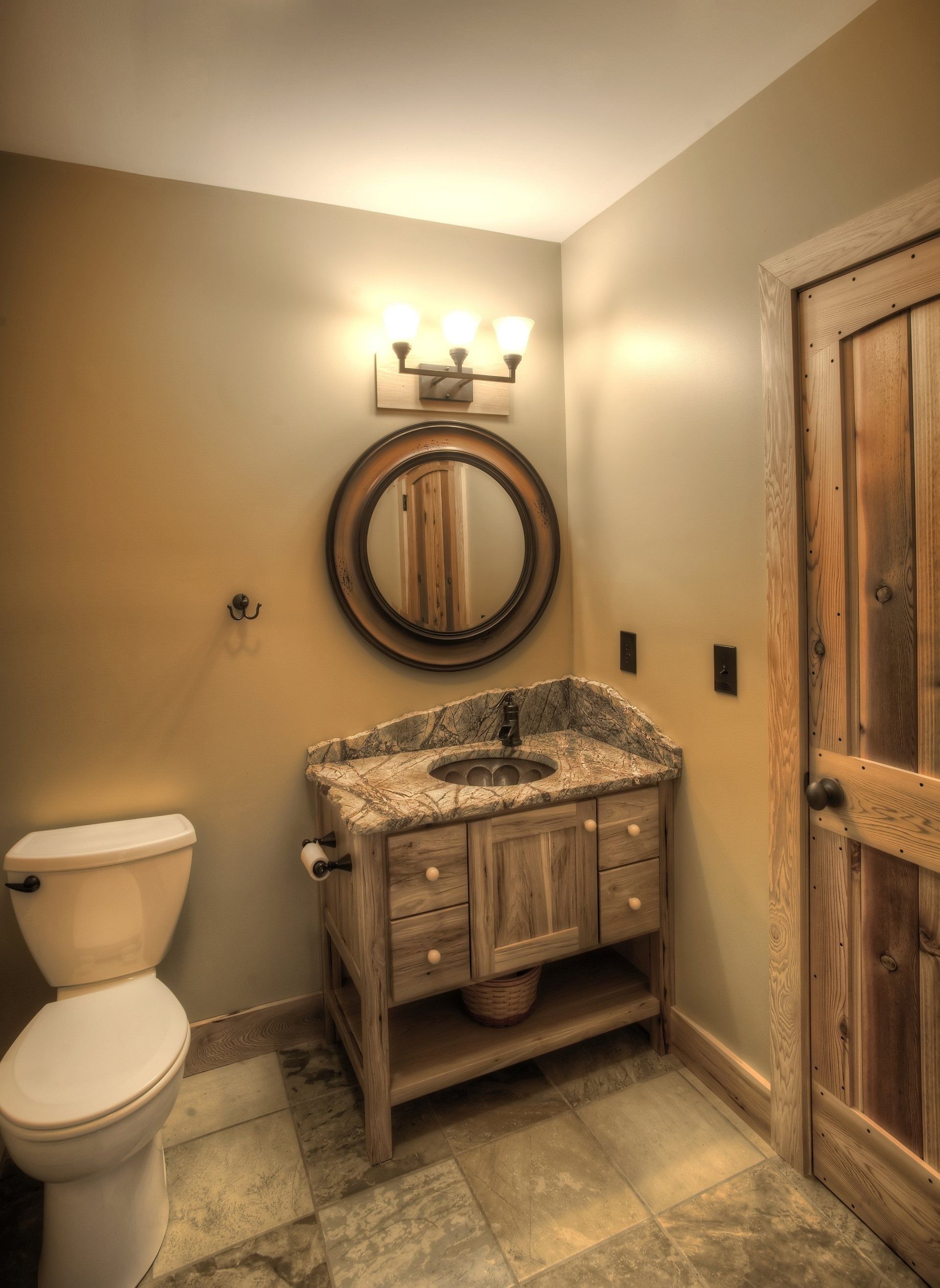 Bathroom with rustic wood vanity, round mirror, and matching door. Beige walls, stone floor.