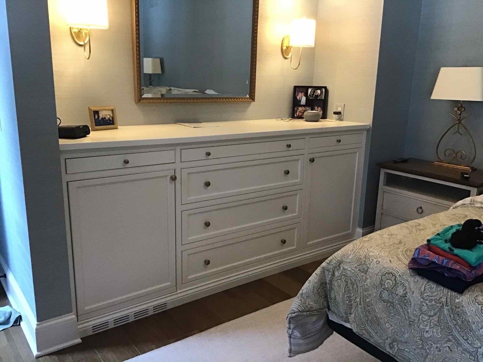White built-in bedroom dresser with mirror, sconces, and nightstand in a room with blue walls and a bed.
