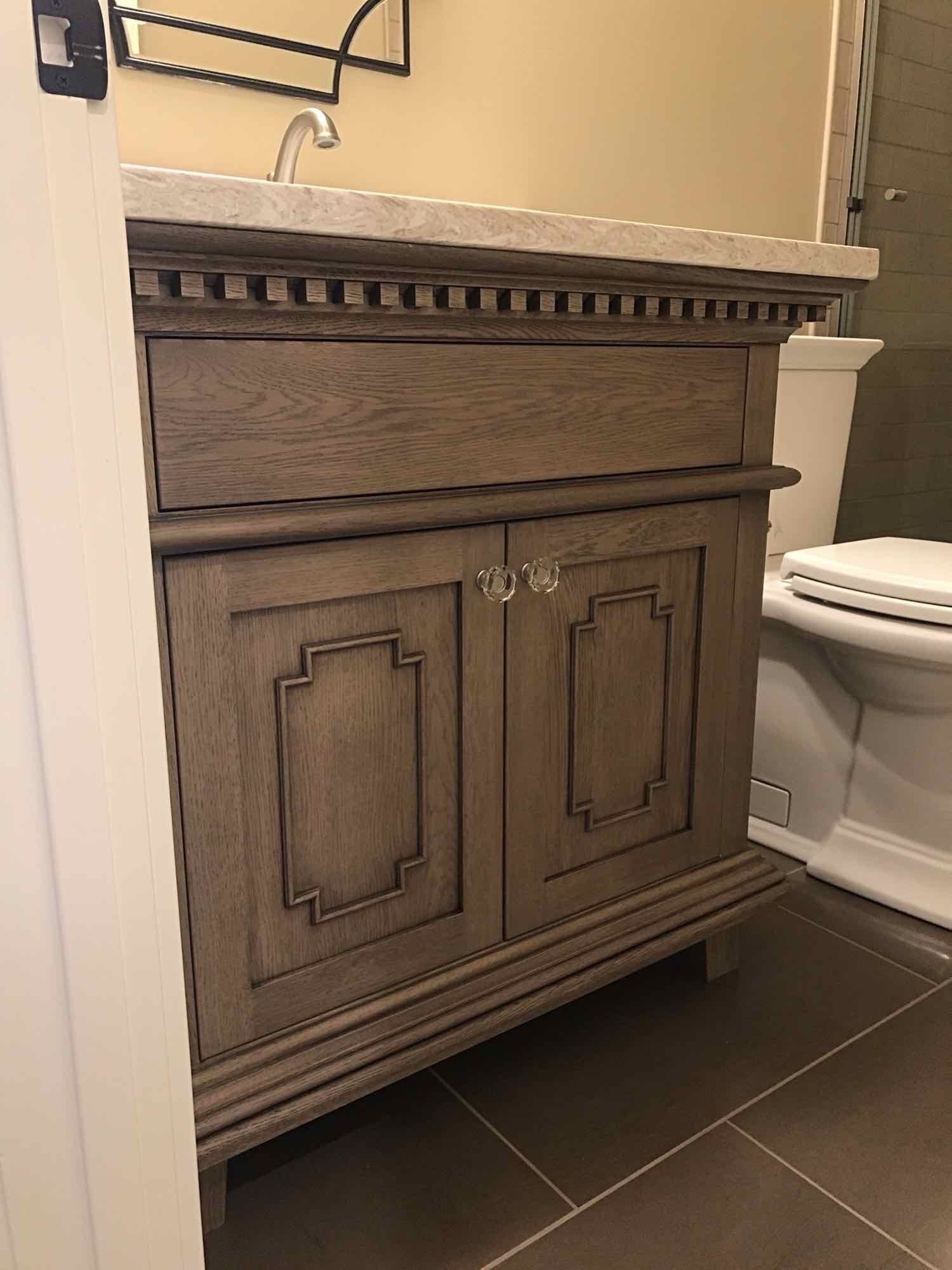Wooden bathroom vanity with white countertop, drawer, and two doors.  A toilet is visible.