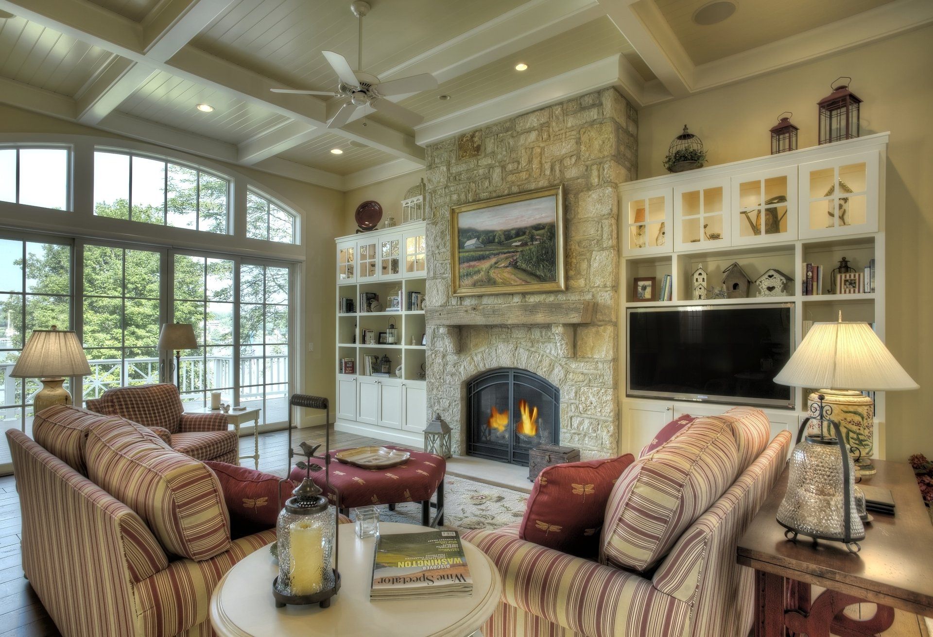 Cozy living room with stone fireplace, built-in shelves, patterned couches, and large windows overlooking a scenic view.