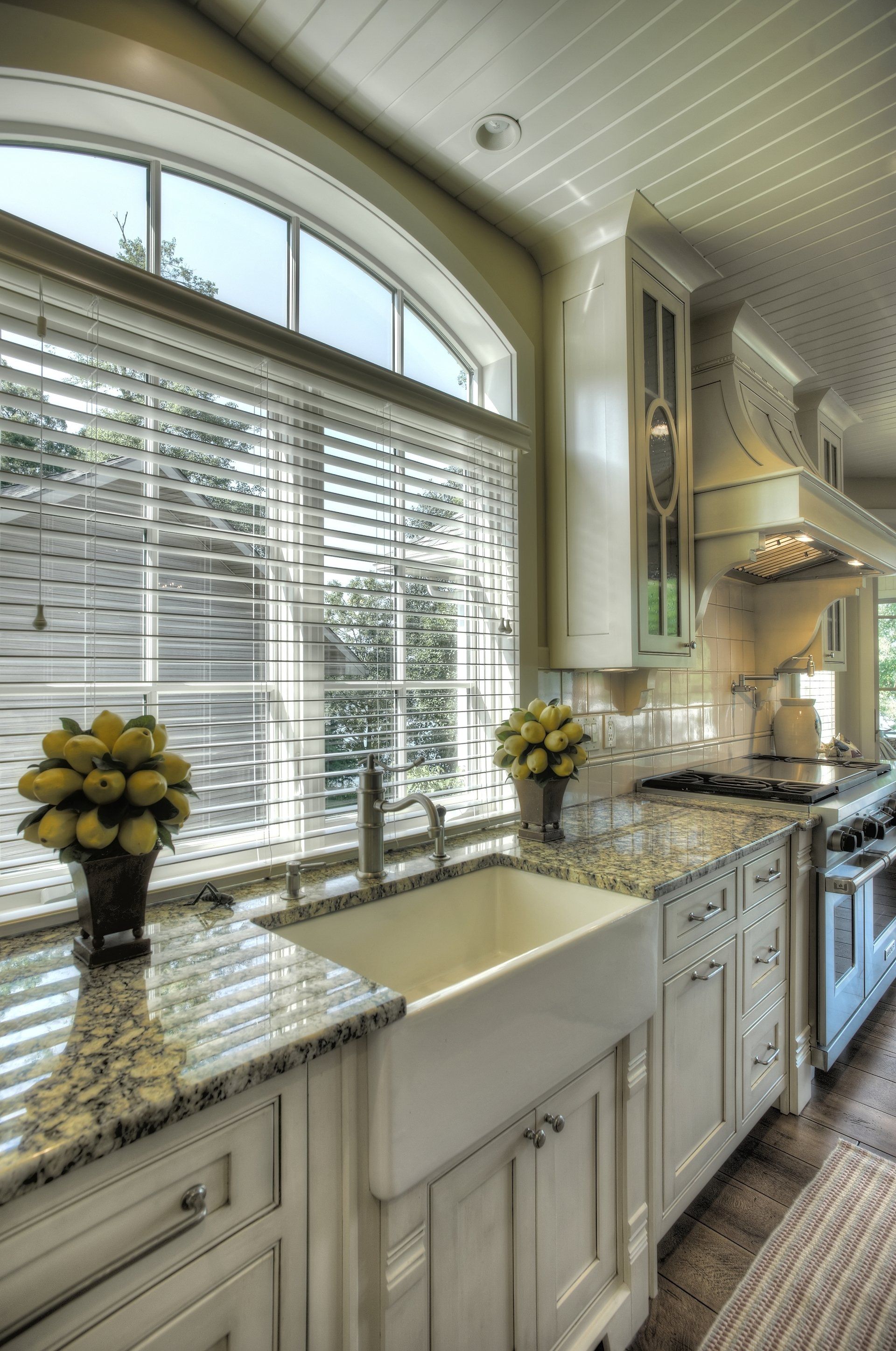 Kitchen with white cabinets, farmhouse sink, granite countertops, arched window with blinds, and lemons.