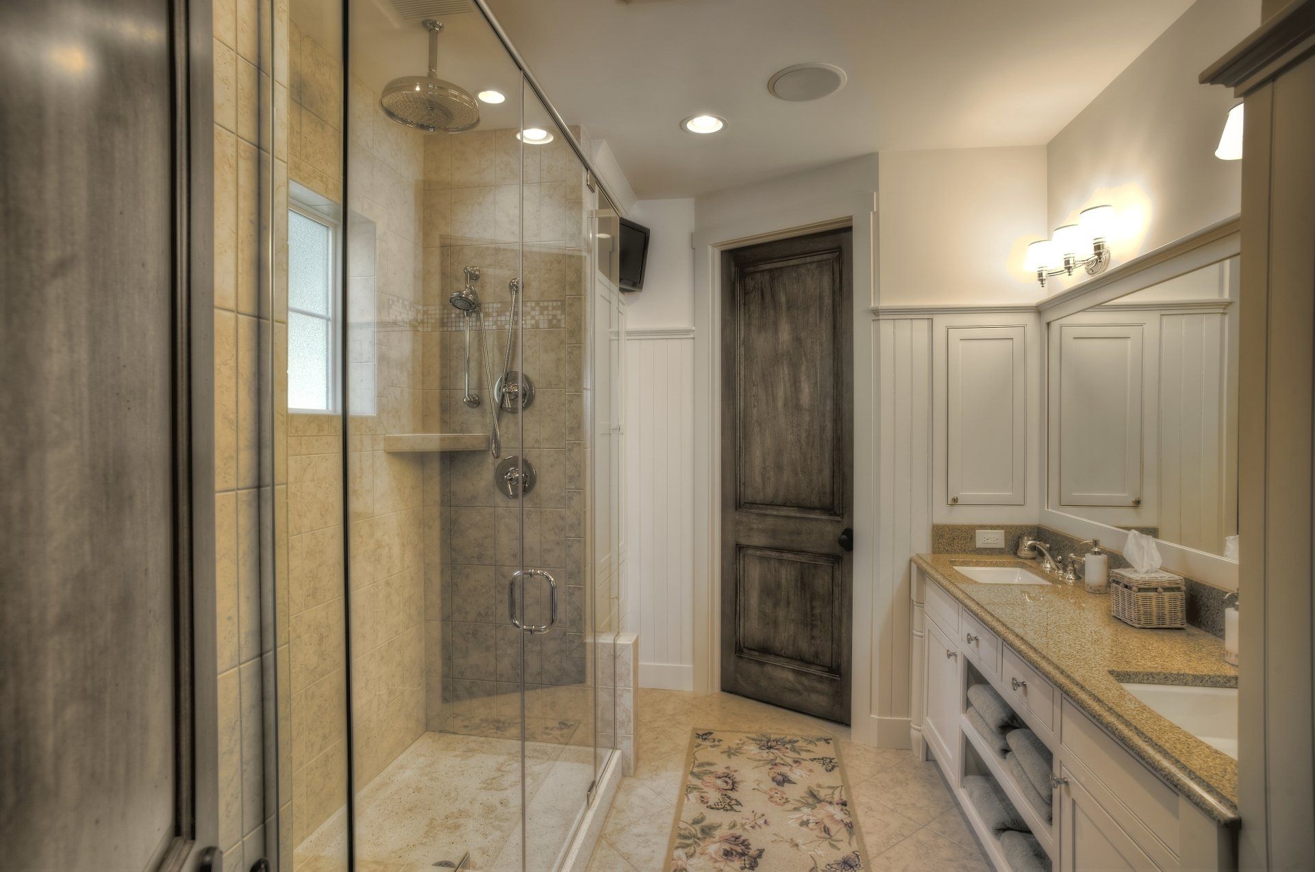 Bathroom with a glass shower, vanity with double sinks, and a dark wooden door.