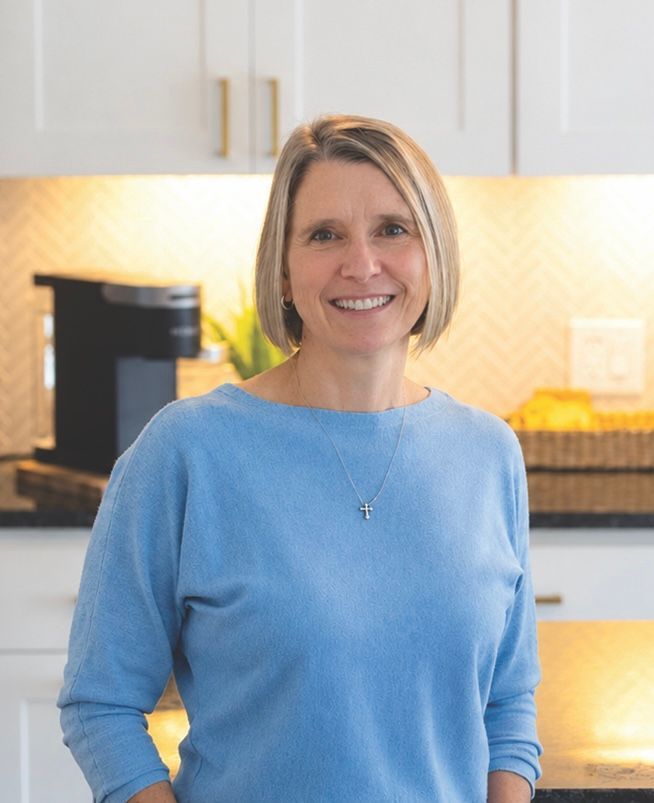 A woman in a blue shirt is standing in a kitchen with her arms crossed and smiling.