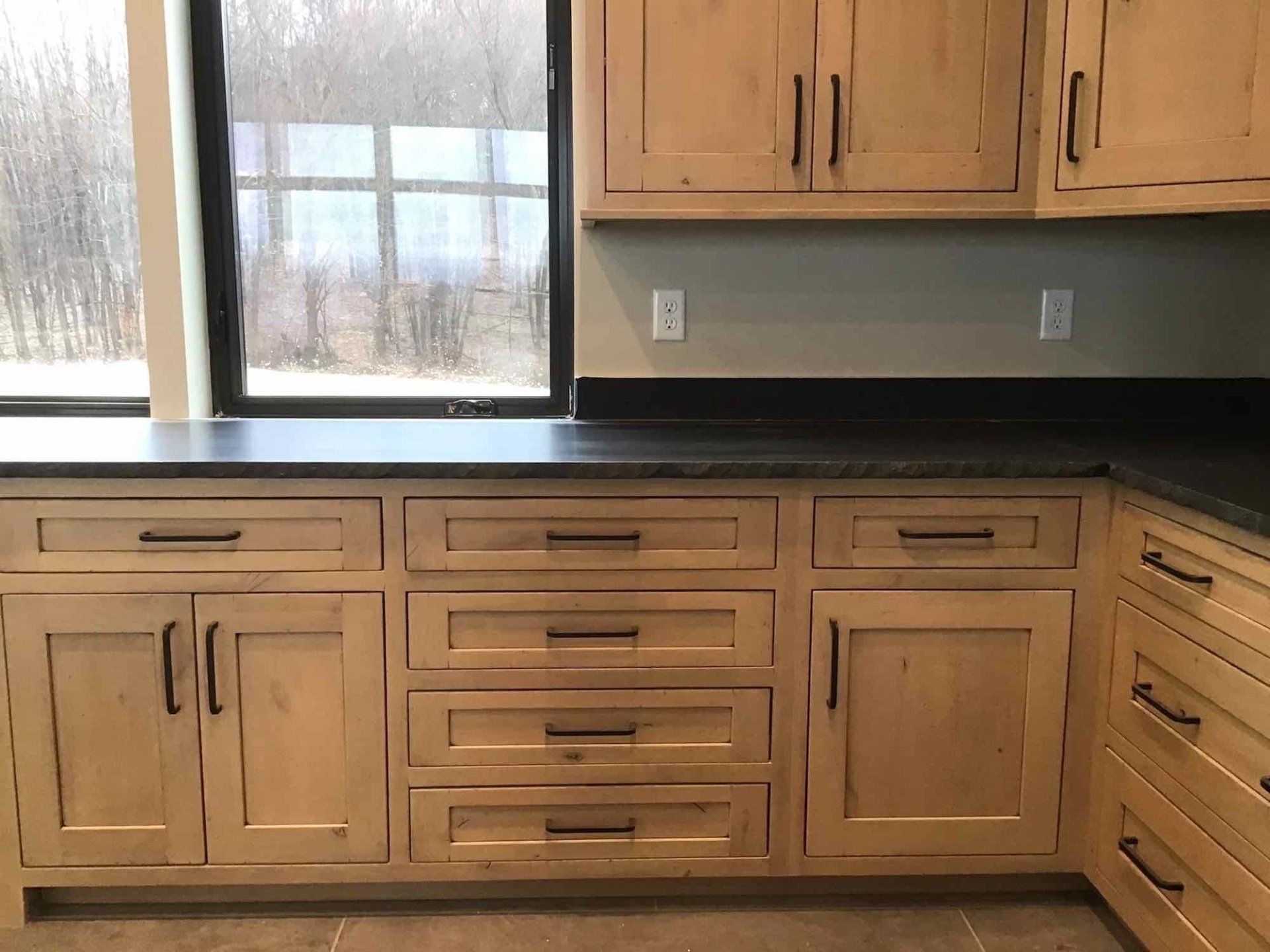 Kitchen cabinets, light wood with black hardware, black countertop, large window in background.
