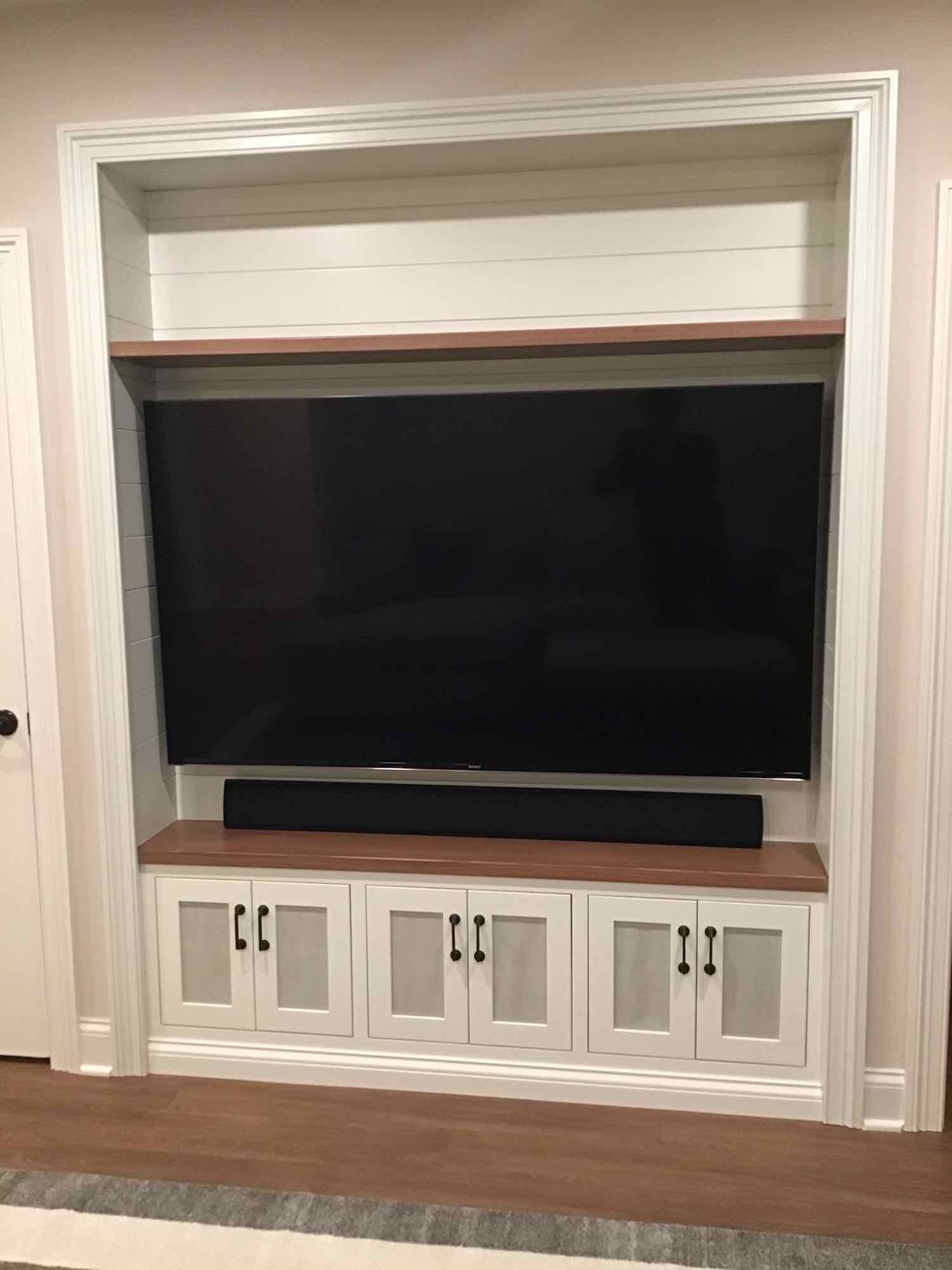 Built-in entertainment center with large TV, soundbar, storage cabinets, and wooden accents within a white frame.