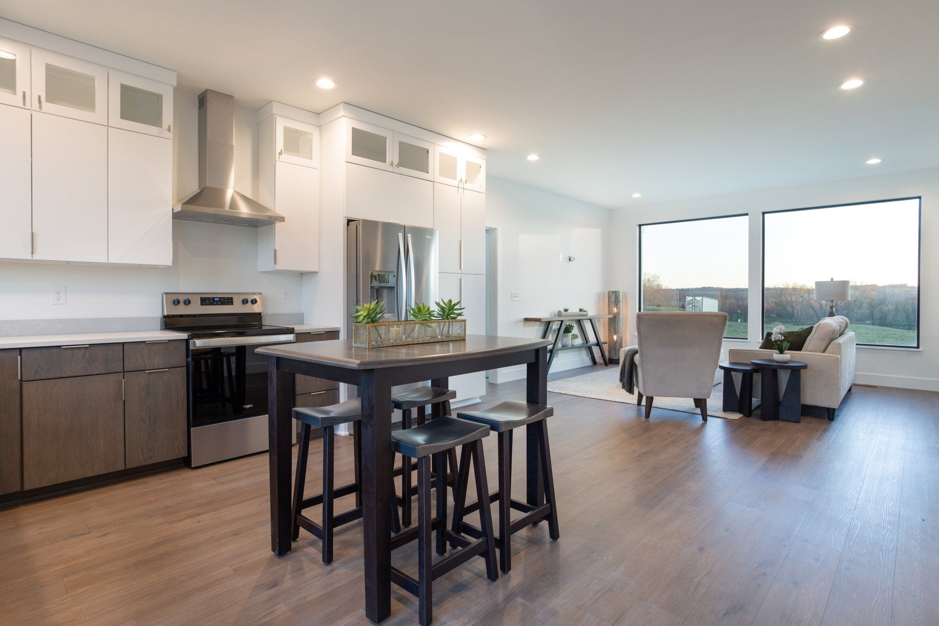 Modern kitchen with dark wood floors, island, and large window overlooking a landscape.