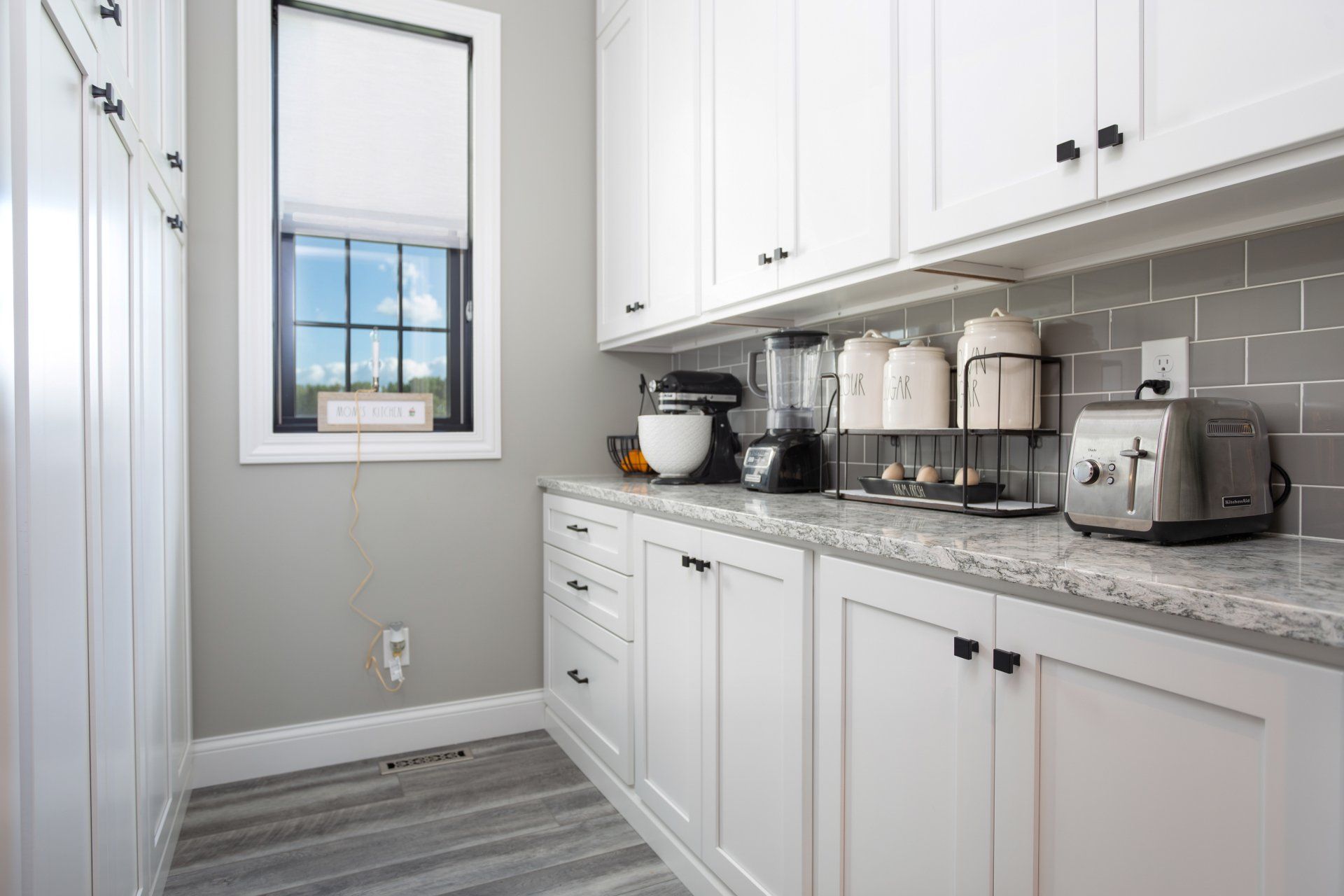 White pantry with appliances, cabinets, countertop, and window with a dark frame and blinds.