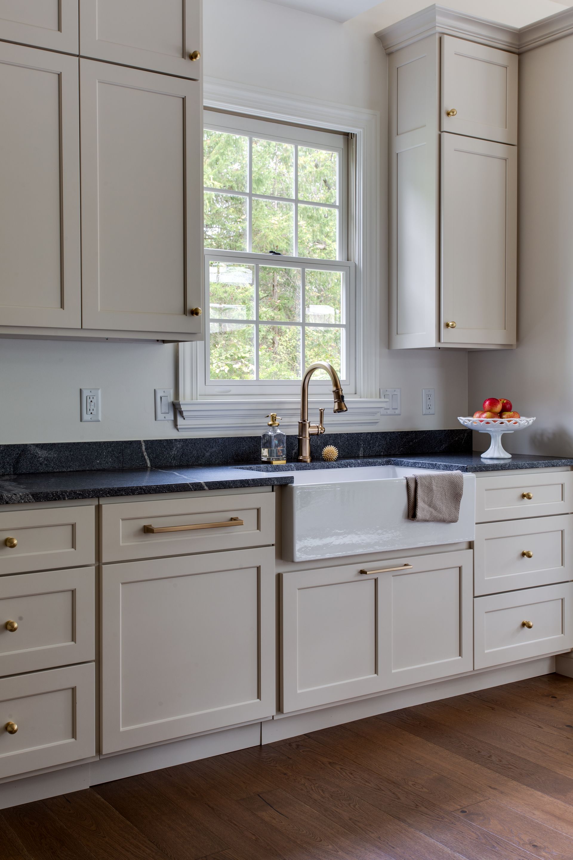 Beige kitchen cabinets surround a white farmhouse sink beneath a window with a dark countertop and gold hardware.